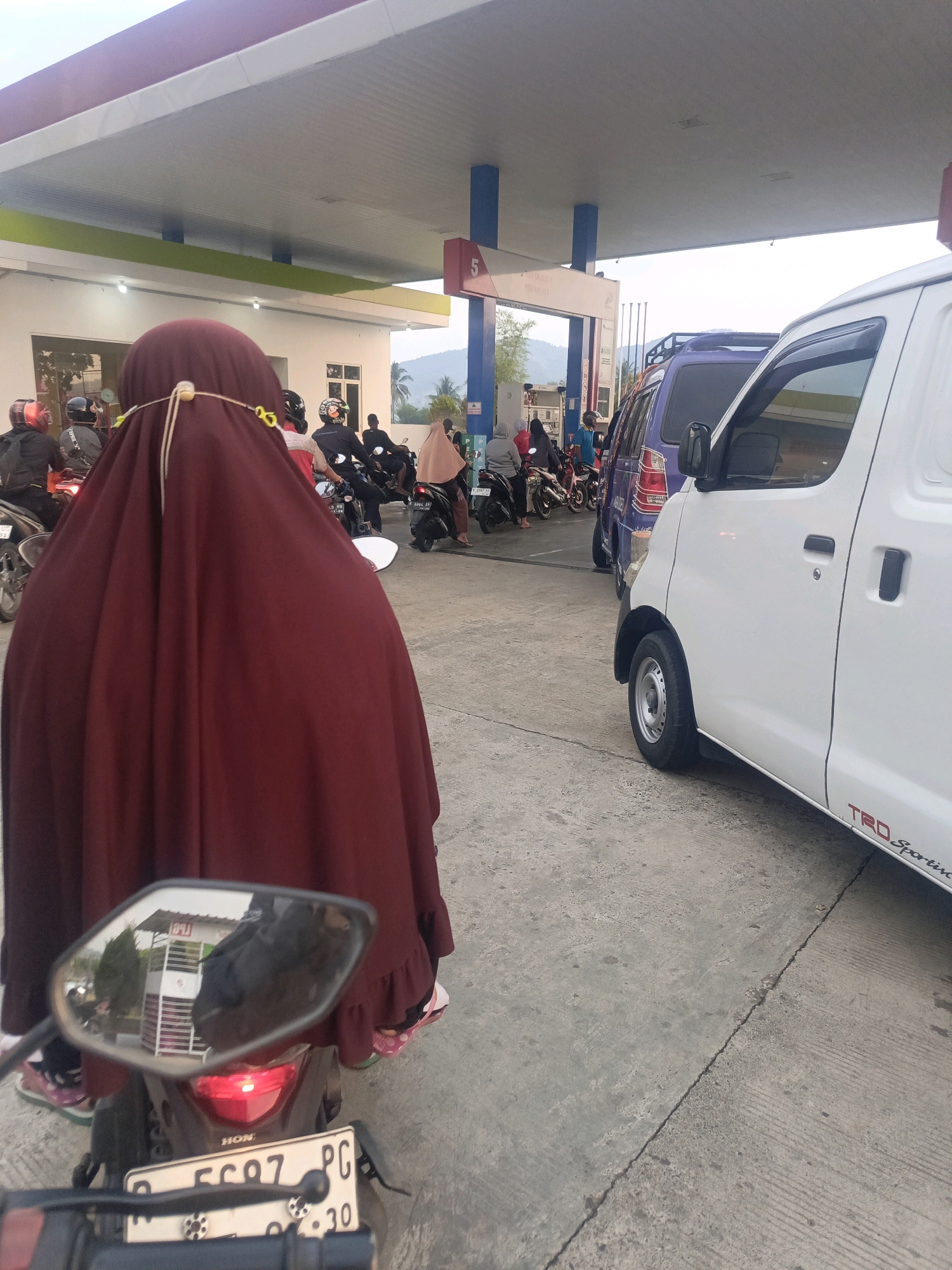The image shows a queue of motorcycles and cars at a gas station in the city of Pegalongan, Indonesia. A person wearing a maroon hijab is riding a motorcycle in the foreground, with the rear of the motorcycle and its license plate visible. Behind them, several other motorcycles and two vans are lined up. The gas station has a red and white canopy. The ground is paved and shows some wear. The sky is overcast.