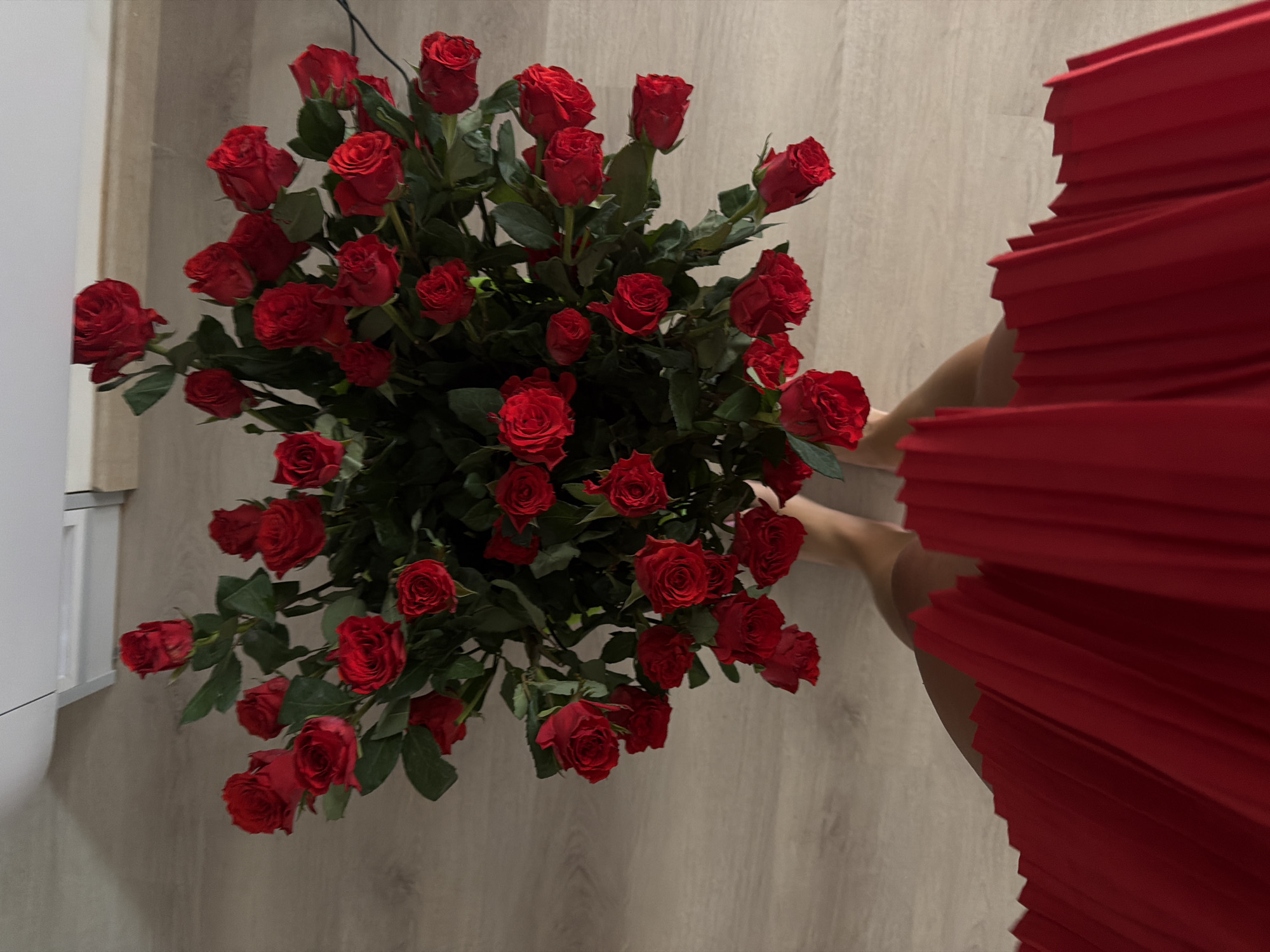 A high-angle shot features a large bouquet of red roses on a light-colored wooden floor. To the right, partially visible, is a person wearing a red pleated dress. The bouquet is the focal point, displaying numerous red roses with green leaves. Part of a white cabinet or wall is visible on the left side of the frame. The setting suggests an indoor environment.