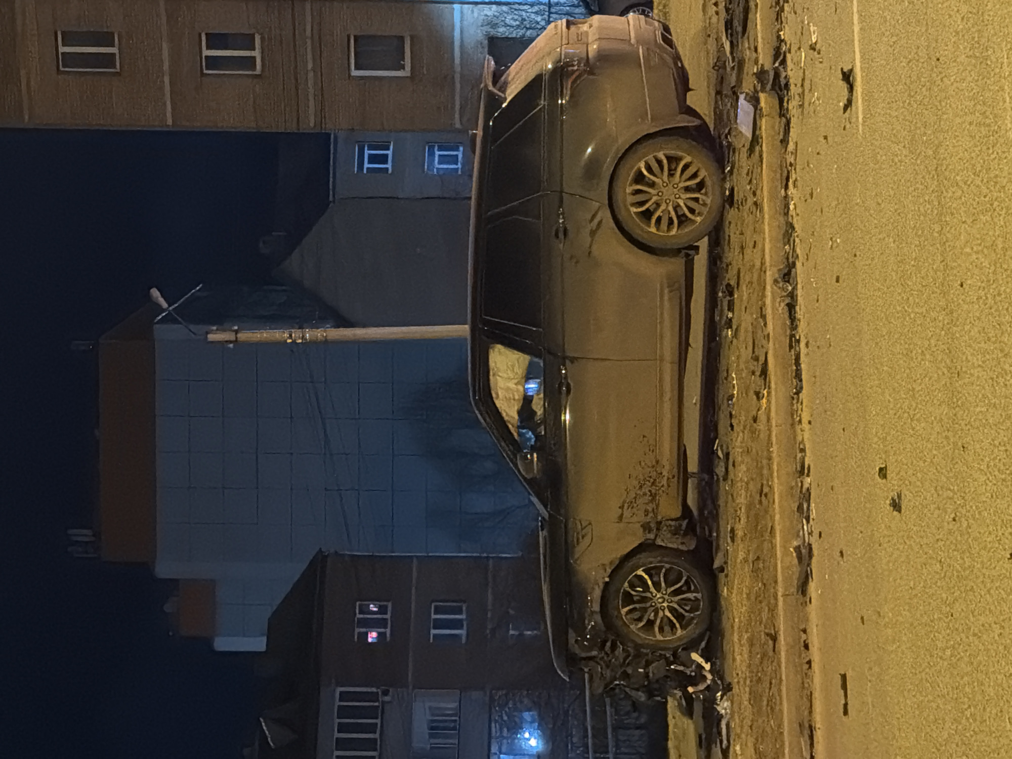 The vertical image, rotated 90 degrees clockwise, captures an urban night scene. A dark-colored SUV, possibly a Range Rover or similar model, is centrally positioned, appearing stationary on the side of a paved road, adjacent to a curb. The vehicle's body is notably dirty, and its front left wheel area shows signs of damage, with debris scattered on the ground beneath it, suggesting the aftermath of an incident. The car features multi-spoke alloy wheels.
In the background, several multi-story buildings, likely residential or commercial, are dimly visible under the dark night sky. Many of their windows are illuminated, confirming the late hour. The architecture of these buildings appears utilitarian. A utility pole or street light is partially visible on the left side of the frame. The immediate foreground and the road surface are illuminated by a warm, yellow artificial light, characteristic of streetlights. No individuals are present, nor is any discernible text visible in the scene. The overall impression is a quiet, late-night urban environment.