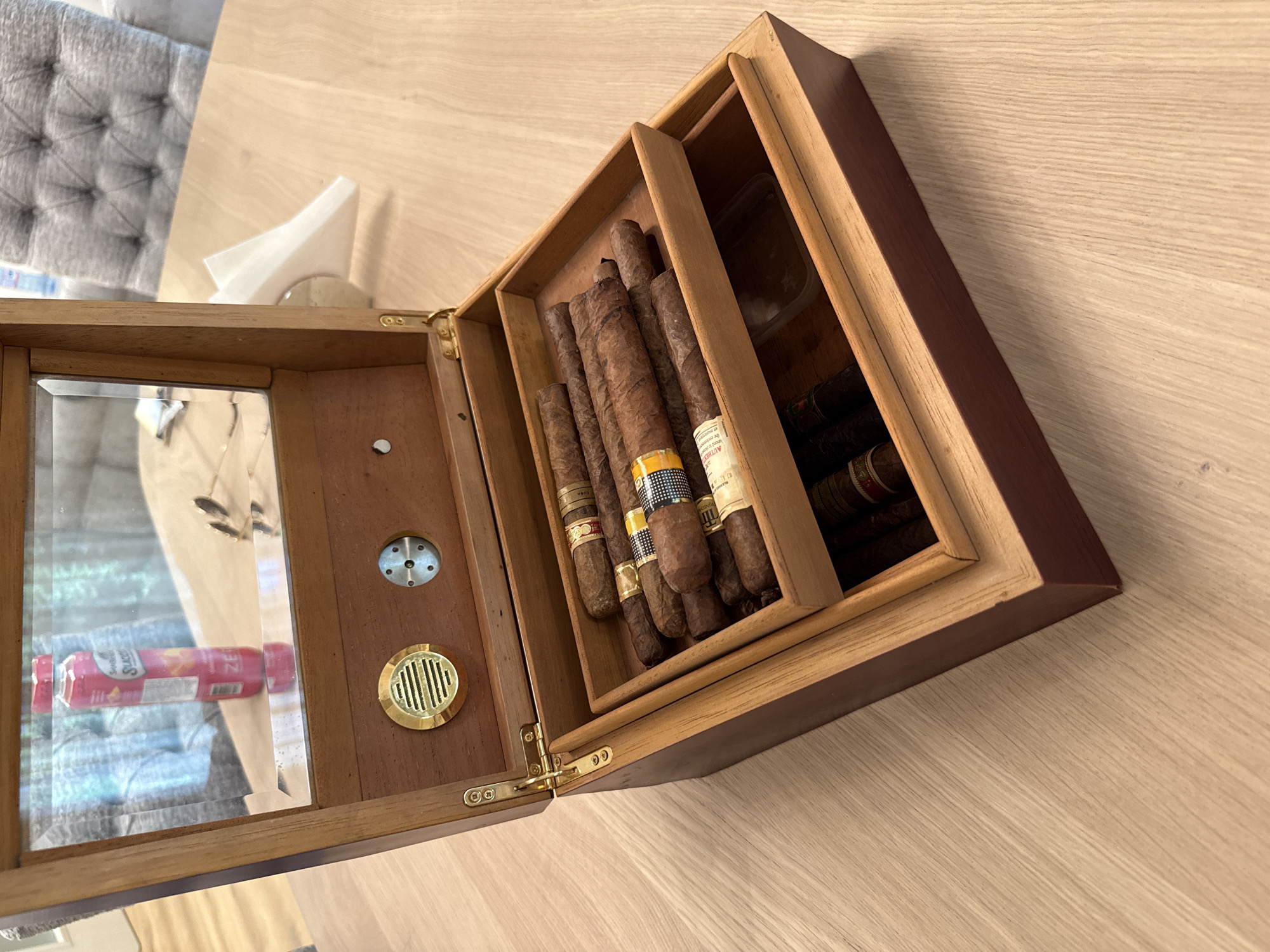 A partially open wooden humidor, holding cigars with Cuban branding, rests on a light-colored wooden surface. The humidor features a glass-covered lid with hygrometer and humidifier elements, and multiple tiers for cigar storage. Some cigars have recognizable branding, including the Cohiba brand. The location cannot be determined from this image.