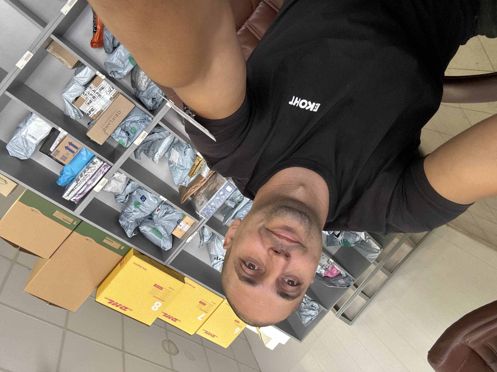 A man is taking a selfie in what appears to be a mail or delivery sorting facility. He is wearing a black t-shirt with a white logo that says "EKOHT". The background consists of grey shelves filled with numerous packages, some in plastic bags and others in cardboard boxes. Several yellow DHL Express boxes are visible on the top shelves. The man is seated in a brown leather chair. The environment suggests an indoor, organized space for handling shipments.