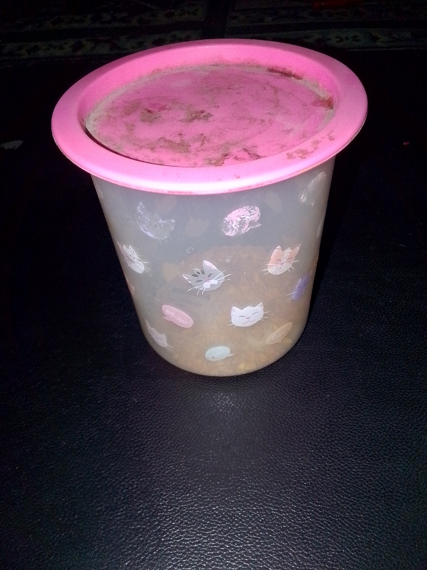 A plastic container with a pink lid, decorated with various cat faces, sits on a dark, textured surface. The container is partially filled with a light brown, granular substance. The lid appears to have some dust or debris on its surface. The background is dark and out of focus, with hints of a patterned textile. The image is taken at a slight angle, looking down at the container.