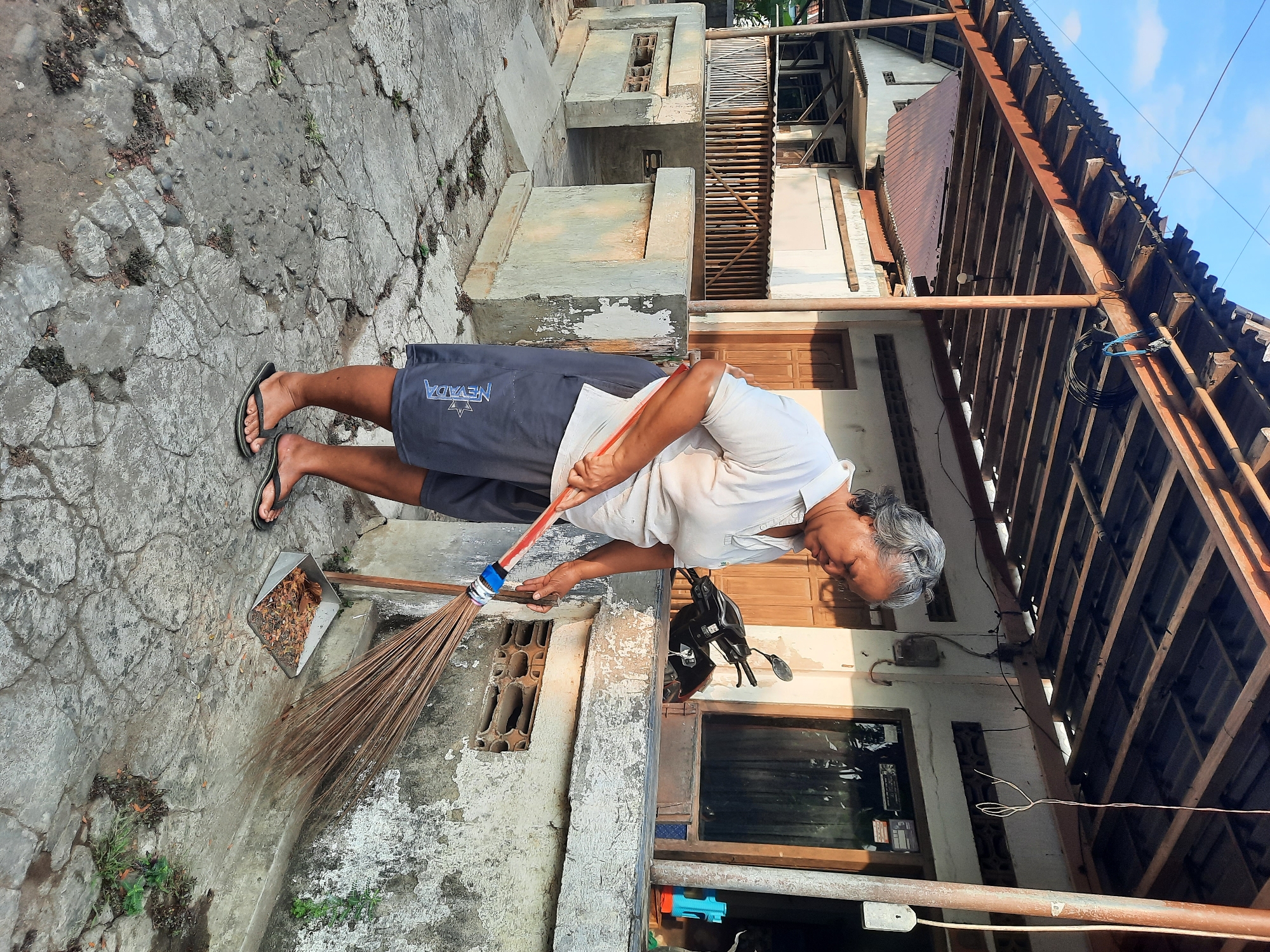 An elderly Indonesian woman with gray hair is sweeping debris with a broom on a cracked concrete patio in front of a house. She is wearing a white polo shirt and dark shorts with the word "NEVADA" printed on them. She is also wearing flip-flops. A dustpan filled with leaves and other debris is placed on the ground next to her. Behind her, a motorcycle is parked next to the house. The house appears to be in disrepair, with peeling paint on the walls and a corrugated metal roof supported by wooden beams. The sky is clear and blue, indicating it is daytime. The scene appears to be a residential area in the city of Andong, Indonesia.