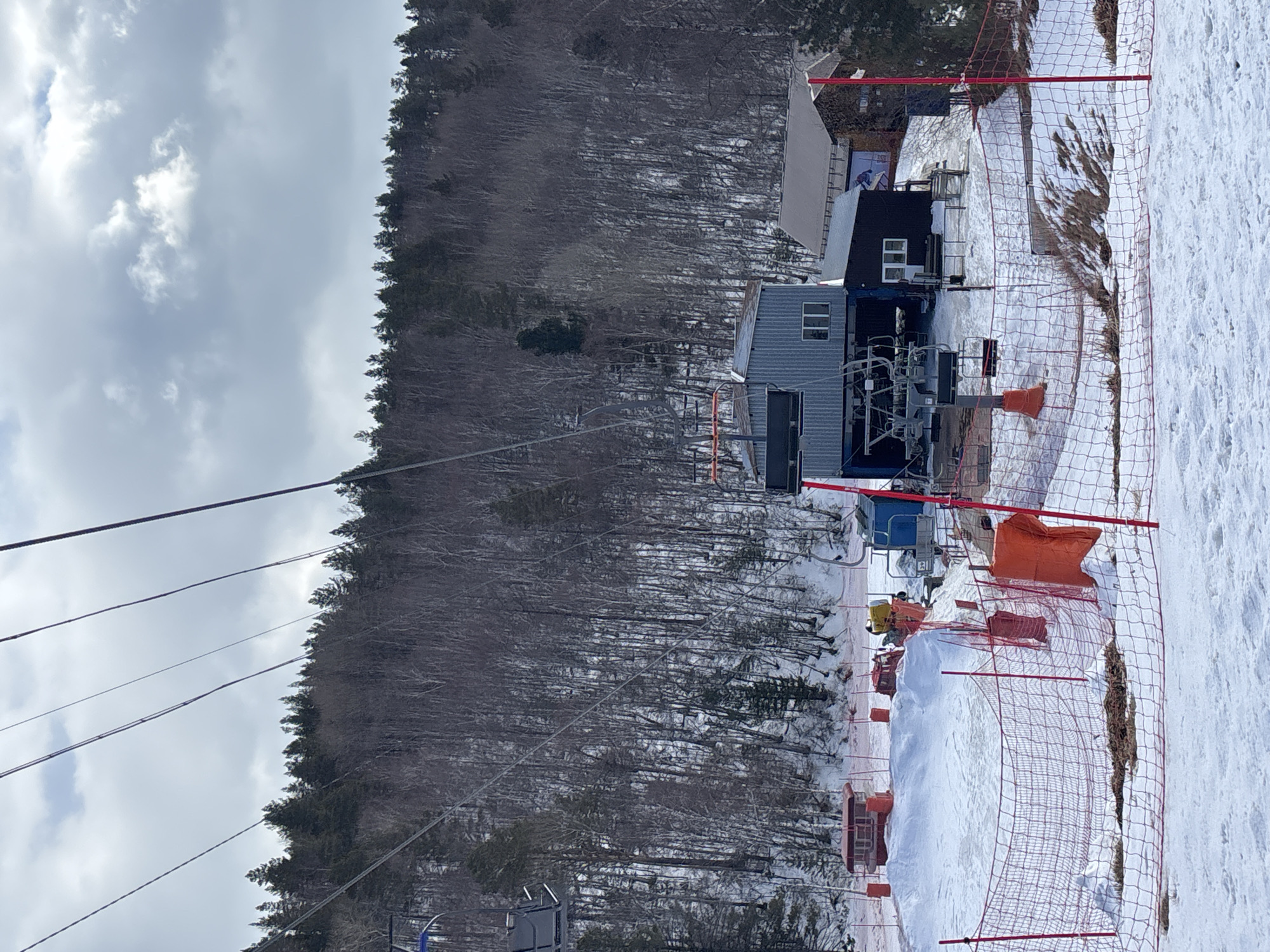 This image depicts a winter ski resort scene in Листвянка, Иркутский region, Russia, on an overcast day. The foreground features snow-covered ground with patches of exposed dry grass and a prominent red safety net on red poles, some with orange padding. In the middle ground, a grey corrugated metal building with a blue base, identified as a ski lift station, stands centrally. Empty ski lift chairs are visible hanging from cables above and near the station. To the right of this building is a smaller, dark-colored wooden building with a gabled roof and a sign displaying a skier. To the left, several red-roofed booths or small structures are positioned on snow mounds. A yellow snow cannon is visible on a snow pile near the central lift station. A single individual in a yellow jacket is partially visible near the snow cannon. Another person in dark attire is obscured near the smaller dark building. The background is dominated by a dense, snow-dusted forest of mixed deciduous (bare) and evergreen trees, extending up a hillside under a cloudy sky. Multiple ski lift cables traverse the upper portion of the frame.