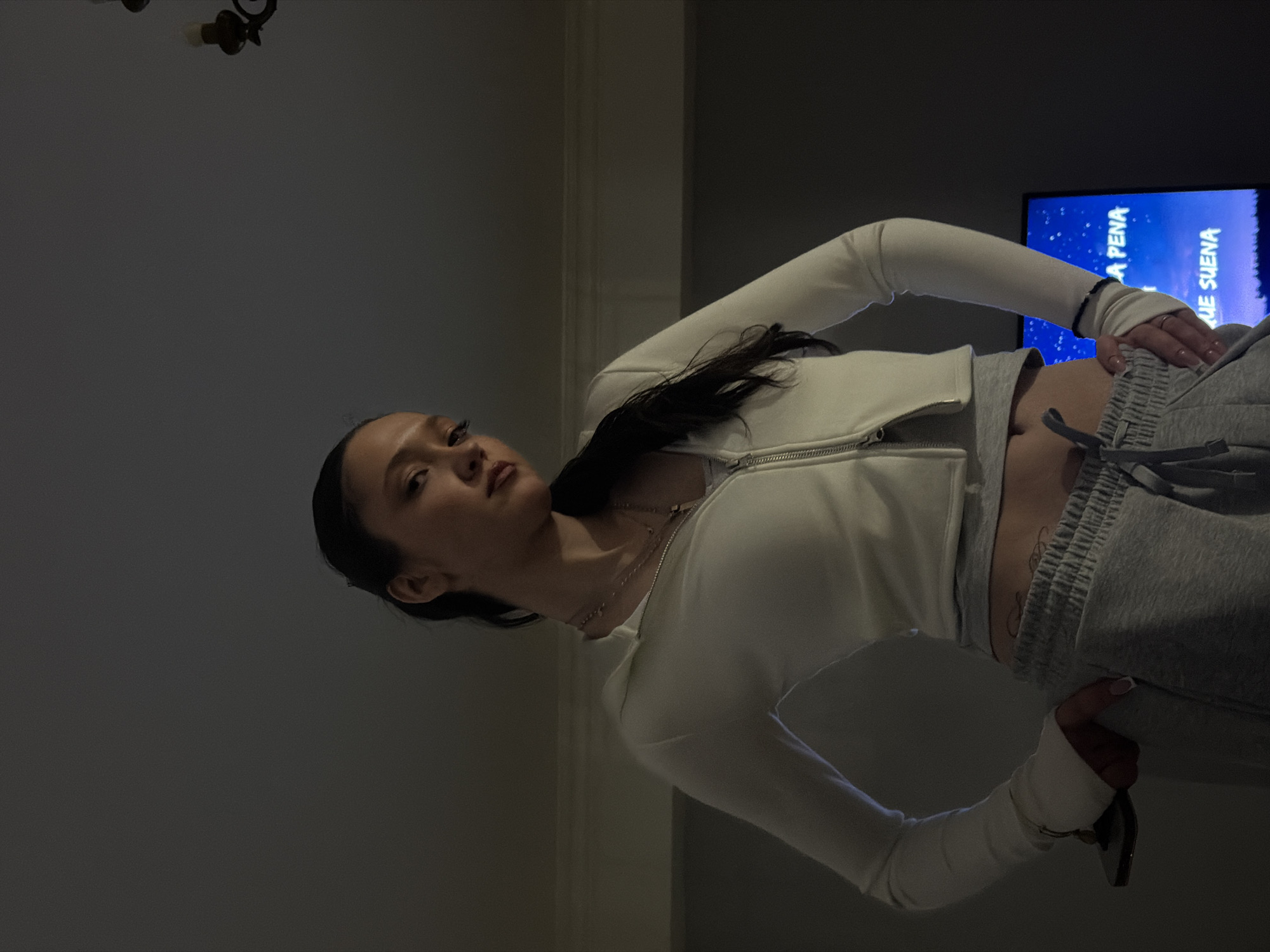 A woman is posing indoors, turned at an angle to the camera. She has dark hair pulled back, and is wearing a white long-sleeved zippered jacket and gray sweatpants. The background includes a TV screen with text in Spanish, and a dark-colored wall. The image does not provide any information about the location in the city of Yerevan, Armenia.