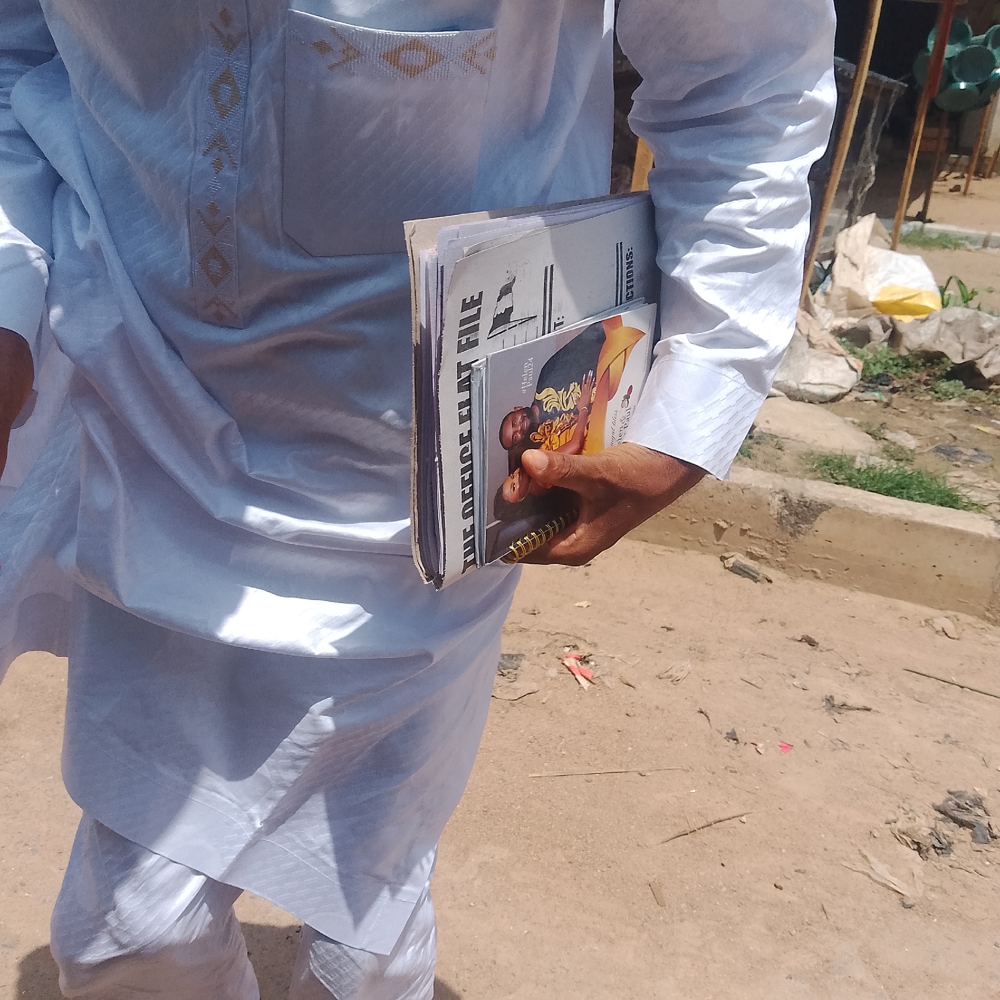In Jalingo, Nigeria, a person wearing a white traditional outfit with gold trim stands on a dirt ground. They are holding a magazine or booklet, which appears to be titled "THE OFFICE FLAT FILE". The person's right hand is visible, holding the booklet. The background shows a dusty environment with some debris and a hint of a green object in the upper right corner. The lighting suggests it is daytime.