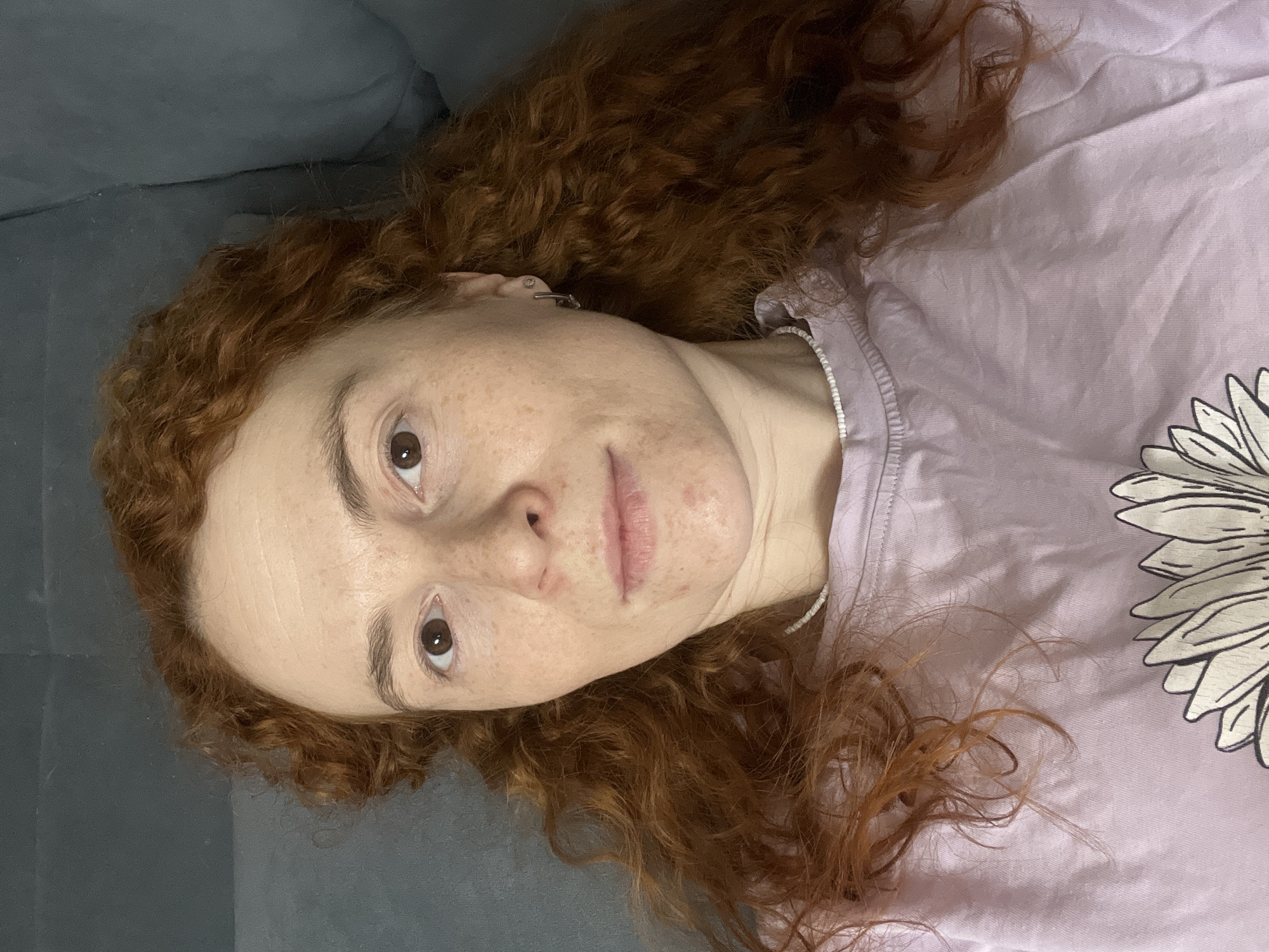A close-up portrait features a person, likely a woman, with fair skin, numerous freckles, and some visible blemishes on her cheeks and chin. She has abundant, curly reddish-brown hair spread around her head. Her dark brown eyes gaze directly at the camera with a neutral, perhaps contemplative, expression.
She is wearing a light purple t-shirt adorned with a large, white flower graphic outlined in black on the right side. A thin, white beaded necklace or chain is visible around her neck, and a small, silvery stud earring is present in her left ear.
The person is lying on a dark grey upholstered surface, which serves as the background, suggesting an indoor setting like a sofa or bed. The lighting is soft and ambient, making the specific time of day or outdoor weather indeterminable. No visible text is present in the image. This media file originates from Chaikovsky, Russia.