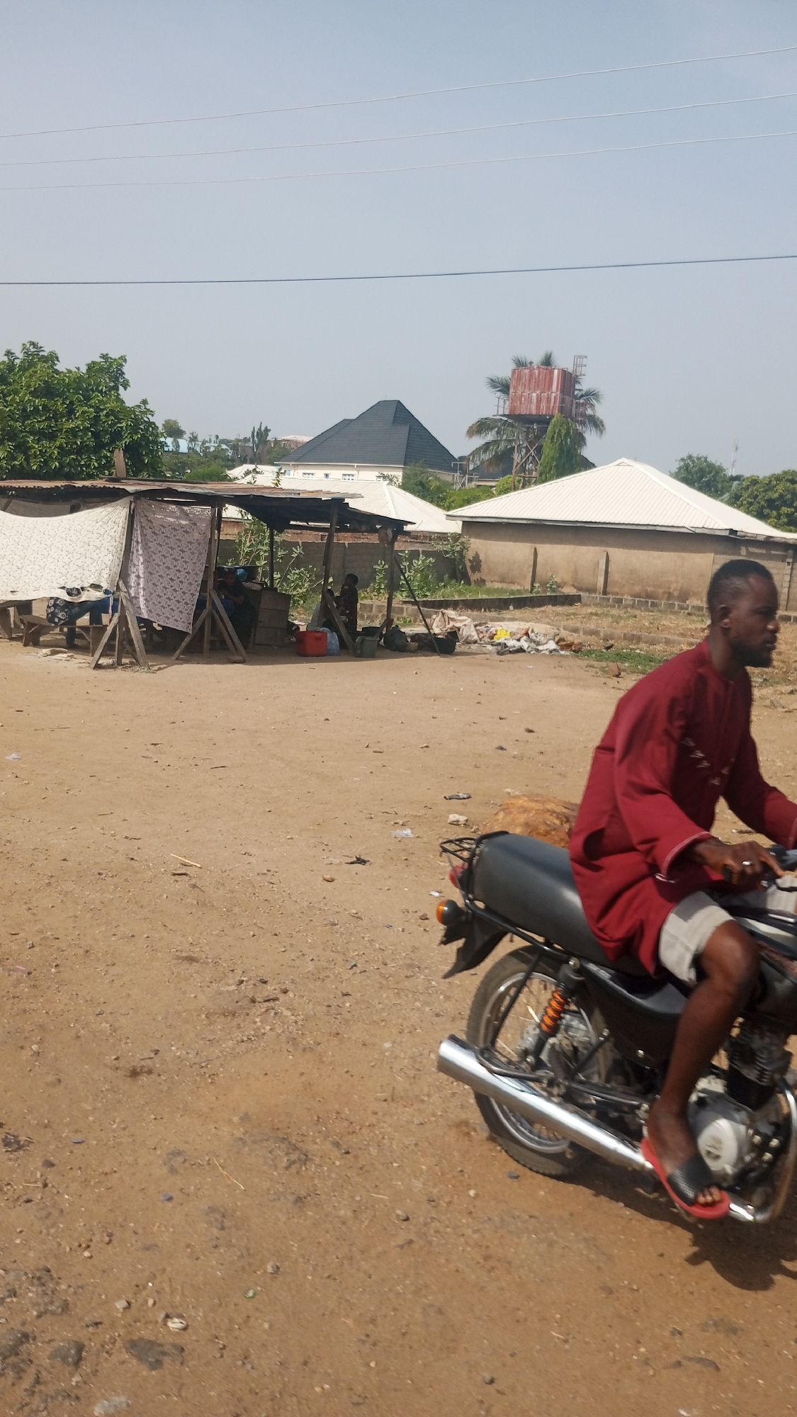 The image depicts a vibrant, sunlit street scene in Lokoja, Nigeria, characterized by a dusty, unpaved road and a mix of traditional and modern structures. The time of day appears to be midday or early afternoon, given the bright, clear sky and strong sunlight.
In the immediate foreground, a man rides a black motorcycle from right to left. He is seen in profile, wearing a long-sleeved, knee-length red garment (possibly a traditional tunic) over light-colored shorts, with black flip-flops on his feet. The motorcycle has a chrome exhaust pipe and a luggage rack at the rear.
To the left, a makeshift stall or market stand is set up on the dirt ground. It's constructed from wooden poles, covered with corrugated metal sheets and patterned fabric panels, offering shade. Several people are visible under the stall, engaged in daily activities, such as sitting on benches or attending to items, including a prominent red cooler.
The background features a blend of buildings and natural elements. Several houses are visible, some with contemporary white corrugated roofs and another with a distinct dark pyramidal tile roof. Further in the distance on the right, a rusty, elevated metal water tank stands on a support structure, surrounded by green trees and palm fronds. Overhead power lines stretch across the wide, pale blue sky. The scene captures the everyday flow of life in a semi-urban or rural Nigerian setting. No visible text is discernible in the image.