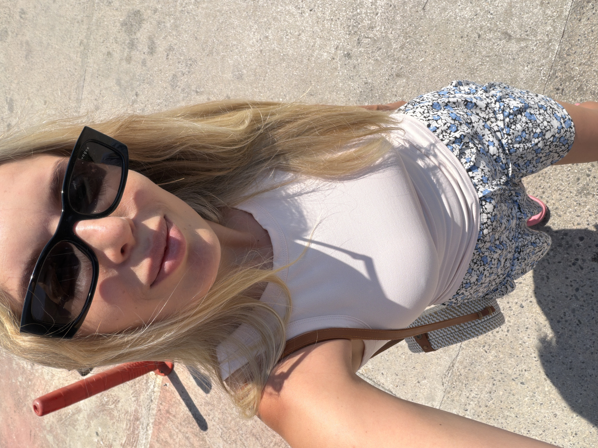 A blonde woman wearing black sunglasses, a white tank top, and floral shorts stands on a concrete surface outdoors, likely a street or promenade, in Budva, Montenegro. A red traffic cone is visible to her left. She is holding a round straw bag with brown leather straps. The sun casts a shadow on the ground.