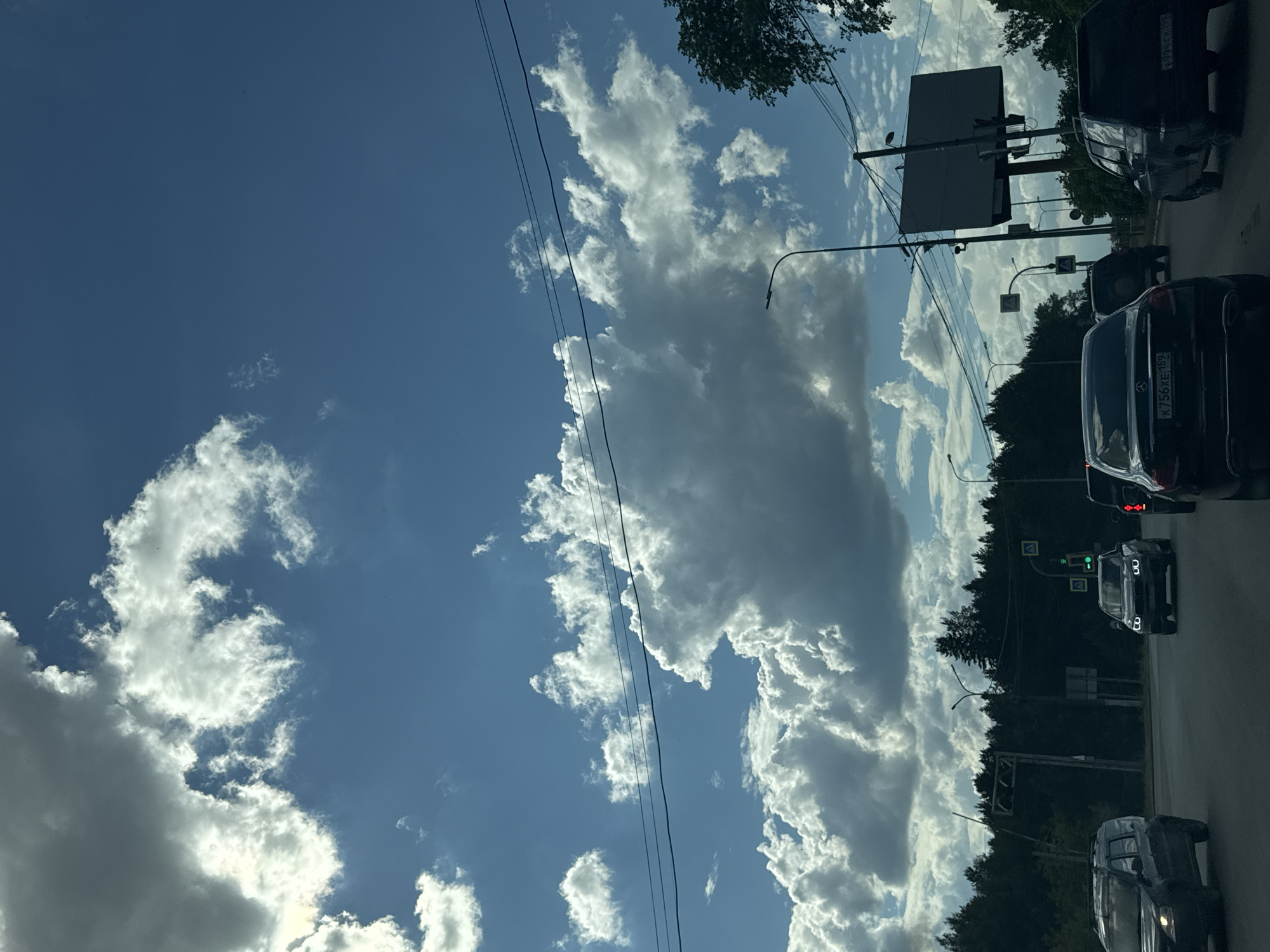 The media file shows a view from a car on a street in Chaikovsky, Russia. The sky is a vibrant blue with dramatic, billowing white and gray clouds, suggesting a partly cloudy day with potential for rain or stormy weather. Several cars are visible on the road, indicating traffic. One black car in the foreground has the license plate K 756 XE 159. There is also a large, dark billboard structure with supports, likely for advertisements, and traffic lights. Trees line the right side of the road, creating a sense of a suburban or semi-rural setting. Overhead power lines are also present, crossing the sky. The lighting suggests it is daytime, possibly late afternoon, given the angle of the light and the shadows. There are no people clearly visible. The overall mood is one of everyday travel under a dynamic sky.