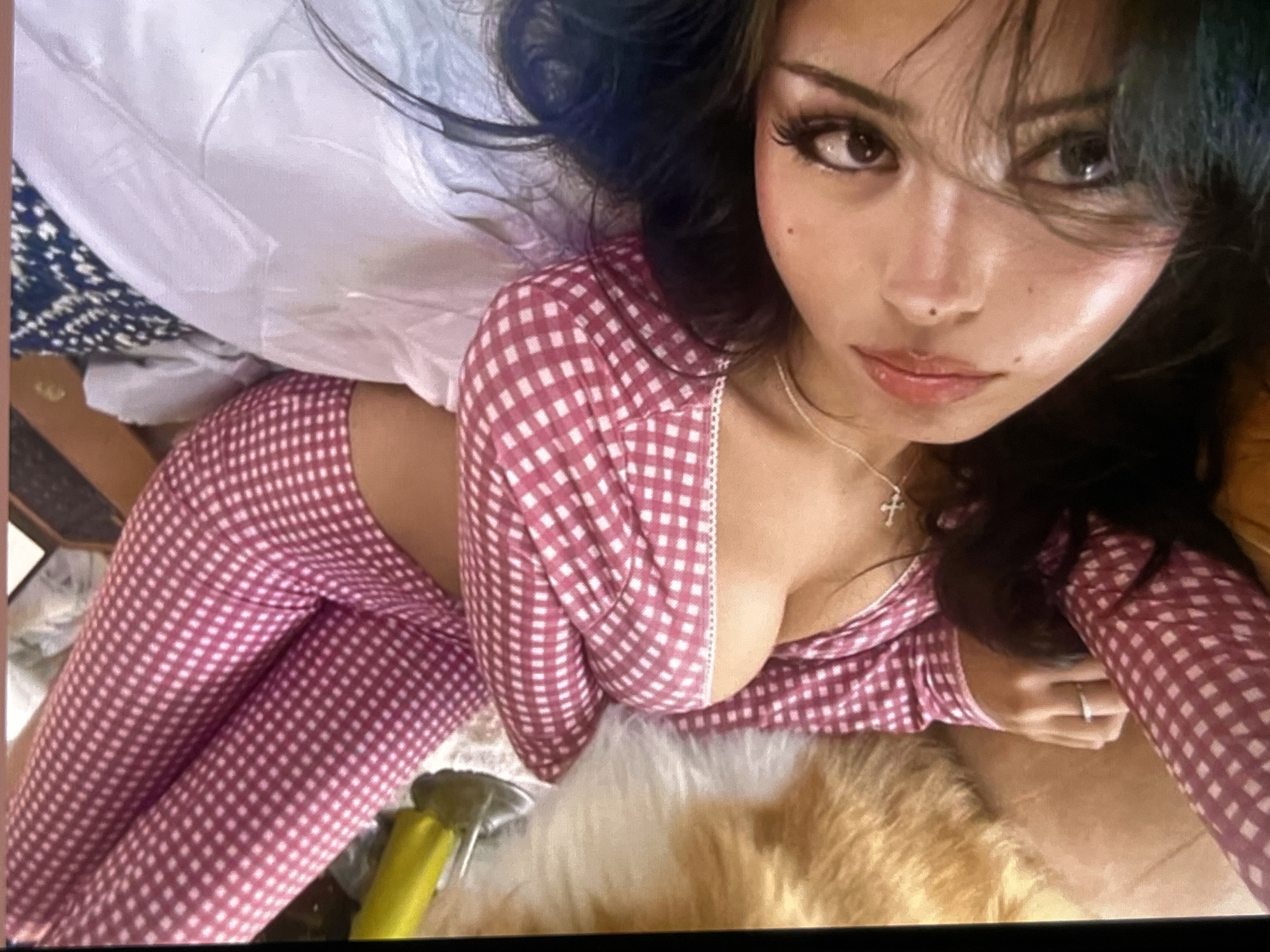 A woman with dark hair and dark eyes lies on a bed with a white comforter and a patterned pillow. She is wearing a pink and white checkered pajama set, which consists of a long-sleeved top and pants. The top has a lace trim along the neckline. She is wearing a ring on her left hand and a cross necklace. A fluffy, light-colored object, possibly a rug or pet, is visible to her left. A yellow object, likely a bottle or handle, is near the top left corner of the image. The overall lighting suggests an indoor setting, possibly a bedroom.