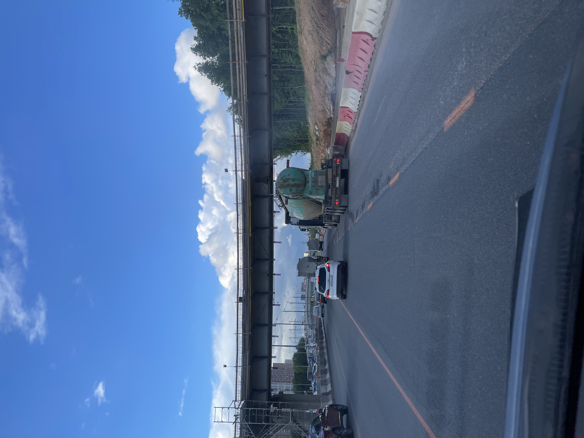 The image shows a view from inside a vehicle traveling on a road, likely in Novodrozhzhino, Russia given the prompt. Above the road, a bridge is visible, possibly under construction, with scaffolding present. The sky is partially obscured by the bridge, revealing blue sky and white clouds. Traffic is present on the road, including a white car directly ahead and a green truck further down the road. A row of red and white road barriers are also along the side of the road. Buildings can be seen in the distance.