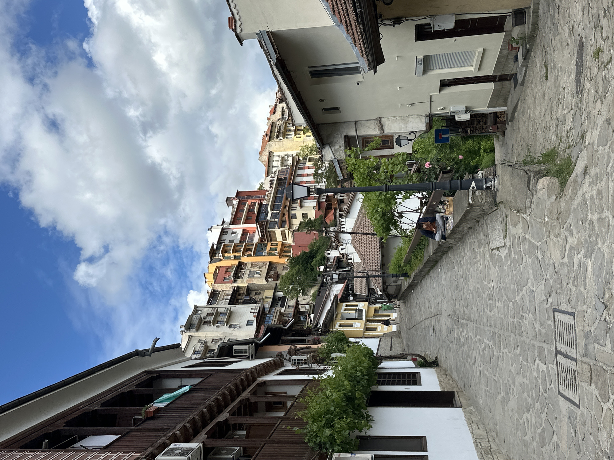 This image depicts a street scene in Veliko Tarnovo, Bulgaria. The view is along a steep, narrow cobblestone street lined with buildings. The buildings are densely packed, with a variety of architectural styles and colors. Some have traditional wooden elements, while others are painted in bright yellows, oranges, and reds.
A light pole stands beside an stone ledge where a man and a woman is sitting. The sky is partly cloudy with bright blue patches.
The focus is on the buildings and the streetscape, which are typical of the historical architecture in the city. The street is made of irregular stone pavement.