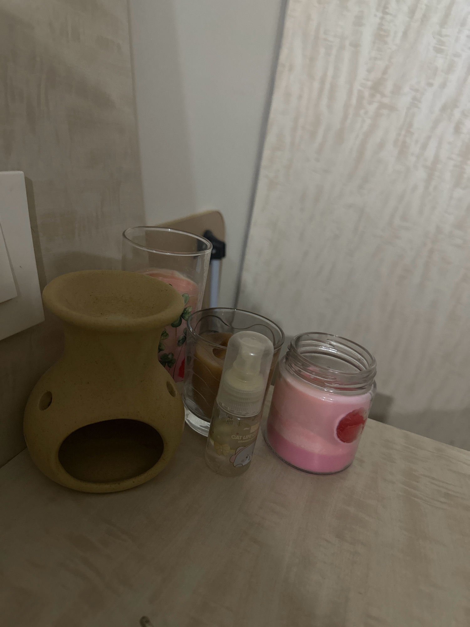 The image displays a collection of decorative items on a wooden surface, likely a shelf or table, in an indoor setting. To the left, a light-colored ceramic oil burner with a hollow opening at the bottom and two circular indentations on the sides is prominently featured. Behind the oil burner are two clear glass containers. The container on the left holds a pink candle with a green floral pattern. The container on the right contains a brown candle. In front of these candles and to the right of the oil burner is a small, clear spray bottle with a white cap, labeled "CAT LIFE STRAWBERRY." To the right of the spray bottle is a clear glass jar filled with a pink layered candle, adorned with a red circular object embedded within the wax. The background consists of a textured wall with subtle vertical lines. The overall impression is one of a curated display of home decor items. The scene is located in Lalitpur, Nepal.