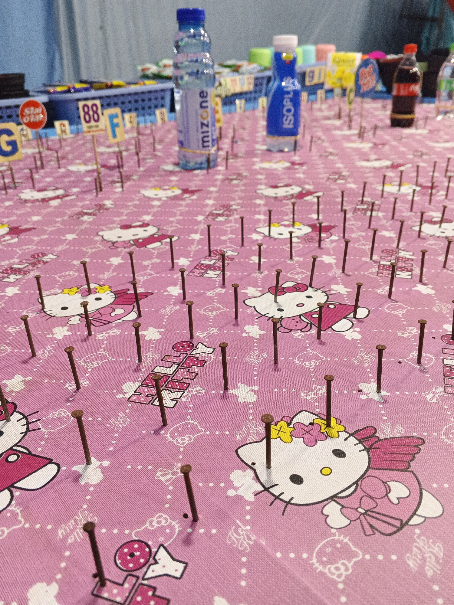 The image depicts a close-up view of a pink tablecloth with a Hello Kitty pattern, upon which numerous small, brown nails are embedded. The nails are arranged in a grid-like pattern, with some placed directly over the Hello Kitty characters. In the background, several bottles of drinks are visible, including a bottle labeled "mizone" and another blue bottle with "ISOPLUS" printed on it. There are also small signs with letters and numbers, such as "G", "88", and "F". The setting appears to be an outdoor stall or booth, possibly at a local market or carnival, given the presence of game elements like the nails and the prizes in the background. The lighting suggests it is daytime. The scene is likely from a game stall in the city of Sukabumi, Indonesia, where such activities are common during local festivities or markets. There are no people clearly visible in the image, and no specific actions are being performed.