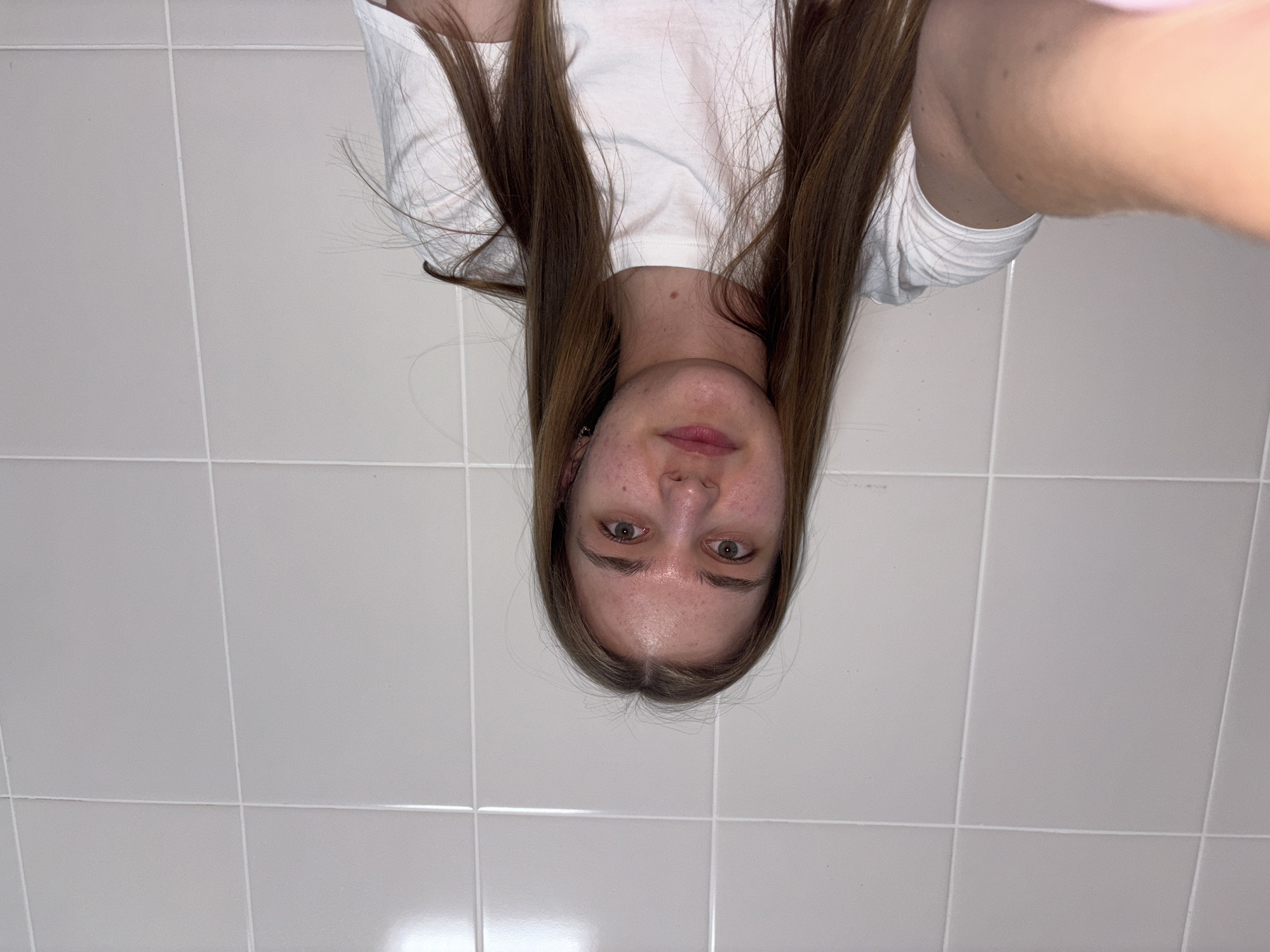 The image shows a person with long brown hair wearing a white t-shirt, likely indoors. The background consists of a tiled wall. No specific location in Perm, Russia can be identified from the image.