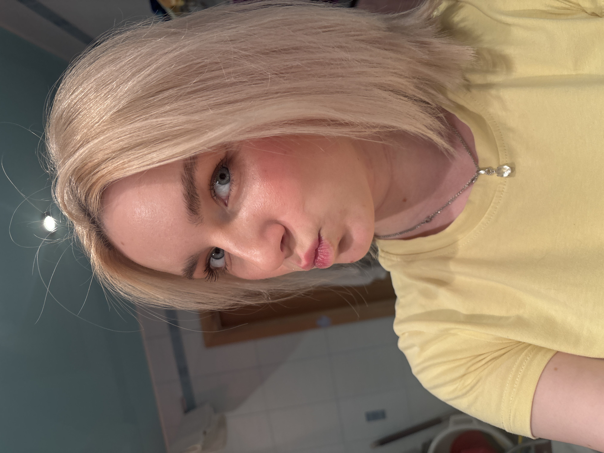 This is a close-up, waist-up, portrait-style, low-angle, indoor photograph of a young woman with blonde hair and light blue eyes. She is wearing a yellow t-shirt and a silver necklace with a small, clear pendant. Her hair is straight and appears to be shoulder-length. The woman is looking directly at the camera with her lips slightly pursed, giving her a playful or pouting expression. Her skin appears fair and flushed, possibly from blush or lighting. The background is out of focus but appears to be a bathroom or similar domestic setting, with tiled walls and what looks like cabinetry. The lighting is artificial and appears to be coming from above. The overall mood is casual and selfie-like. There is no visible text in the image.