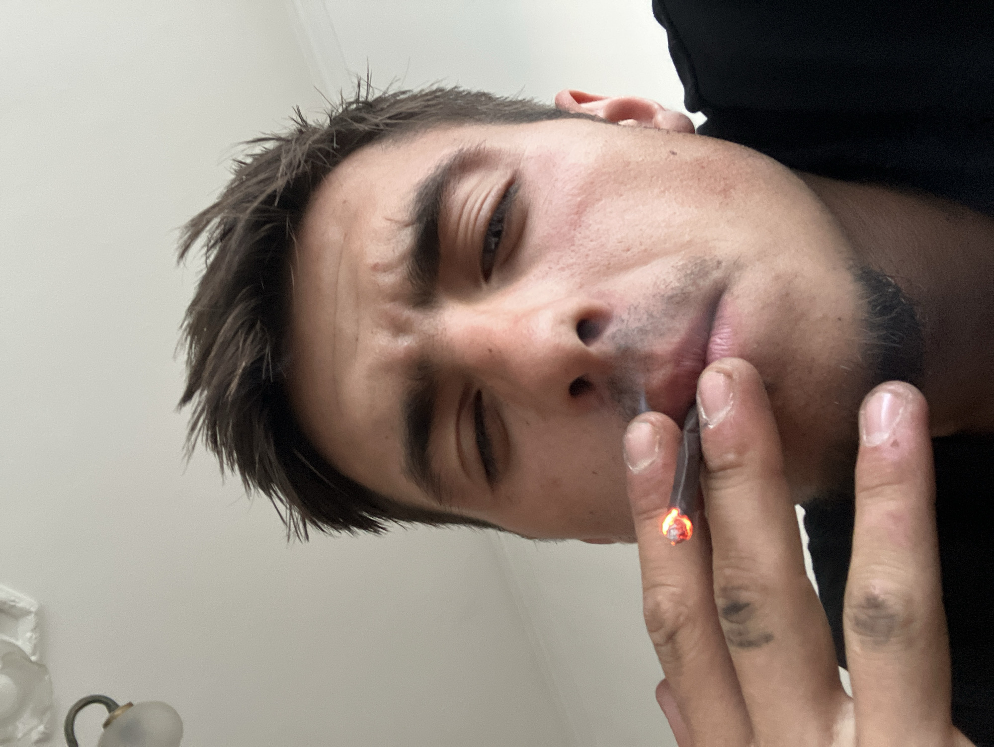 An overhead shot captures a young man from a low angle, looking down towards the camera with his eyes partially closed. He is holding a lit cigarette or joint between the fingers of his right hand, bringing it towards his mouth. The tip of the item is glowing bright orange. His dark hair is slightly dishevelled, and he has light facial hair growth on his chin and upper lip. His skin has some visible pores and minor blemishes.
The man's fingers show visible creases and what appear to be some darker marks or discolouration on the knuckles and between the fingers. He is wearing a dark-colored garment, possibly a t-shirt or hoodie, visible around his neck and shoulder area.
The background is a plain, light-colored wall or ceiling, suggesting an indoor setting. In the bottom left corner of the original image, a white decorative architectural detail, possibly a ceiling molding or part of a light fixture, is partially visible, along with a metallic arm ending in a translucent, frosted glass globe of a light fixture. The lighting is diffuse and ambient, indicating an indoor environment without strong directional light sources. There is no visible text in the image. The scene suggests a private or casual moment of the man smoking indoors in Plovdiv, Bulgaria.