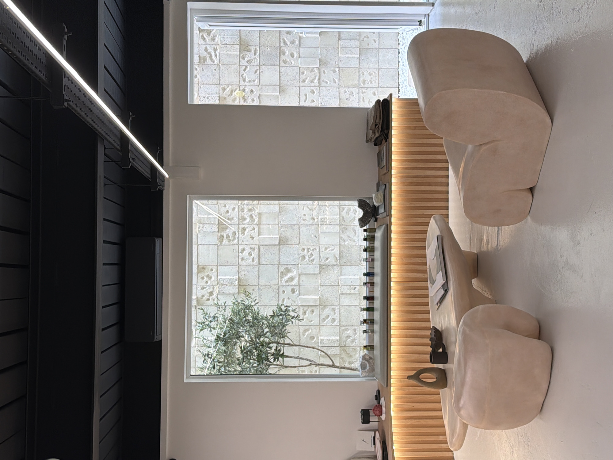 Captured in a brightly lit interior, the view presents a juxtaposed arrangement of textures and materials. The foreground is dominated by two organically shaped, cream-colored armchairs, positioned near a light wooden slat wall. On this wall, a narrow table is adorned with books and decorative objects. Windows frame a view of textured brickwork and the delicate branches of a tree, providing a glimpse of the outside world. A black, slatted ceiling with linear lighting contrasts sharply with the lighter tones of the room. The presence of an air conditioning unit suggests a modern space, although the specific location in London, United Kingdom cannot be definitively determined from the image. There are no people present in the frame.