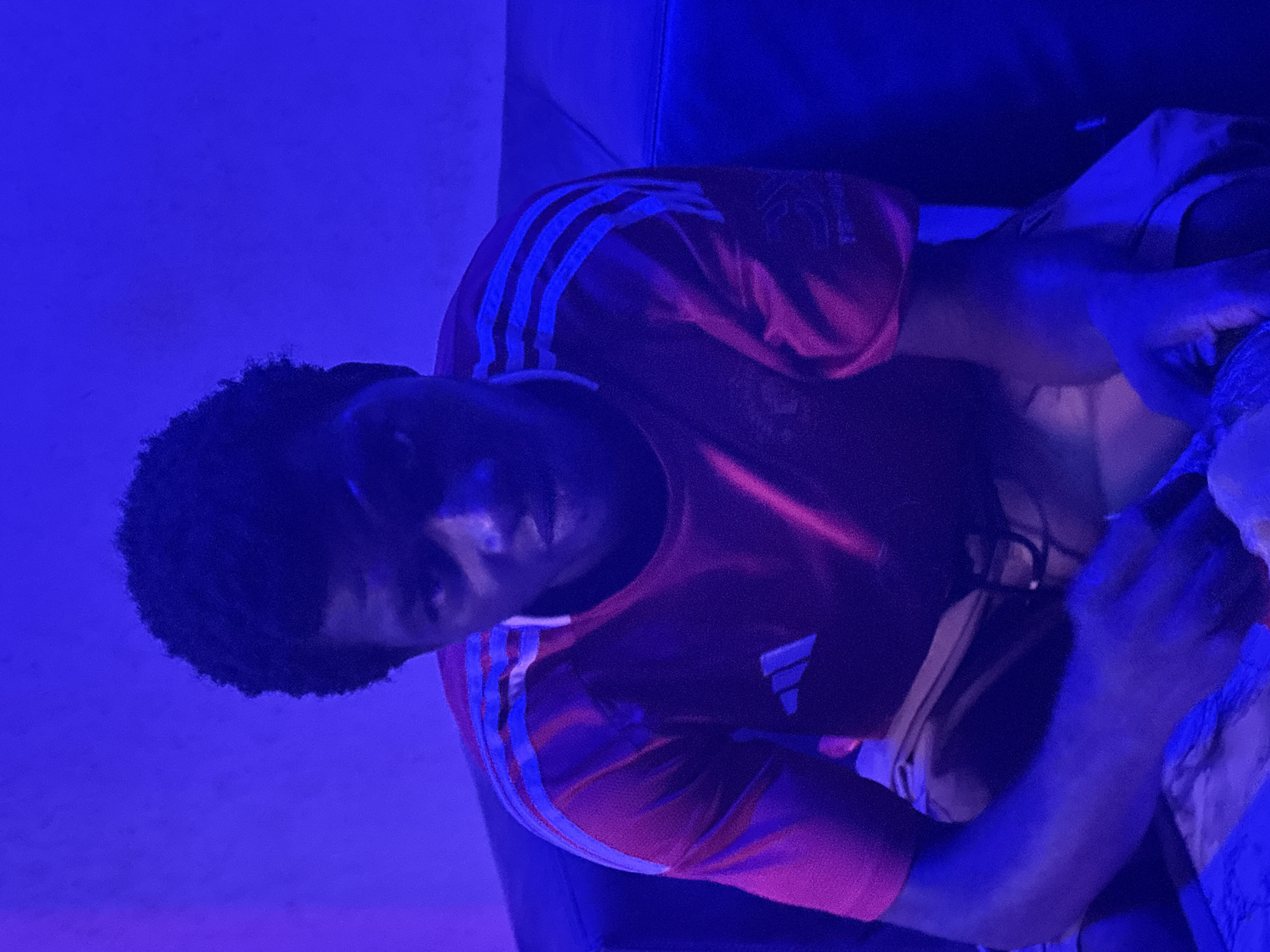 A young Black male is seated on a dark leather couch, facing forward. He has short, dark, curly hair. He is wearing a short-sleeved red shirt with white stripes on the sleeves and an Adidas logo on the left chest. He is also wearing light-colored shorts. The environment is dimly lit with a strong blue light, creating a purplish hue on the subject and surroundings. The background appears to be a wall. The overall impression is that the image was taken indoors, possibly in a residential setting in Ikot Ekpene, Nigeria.