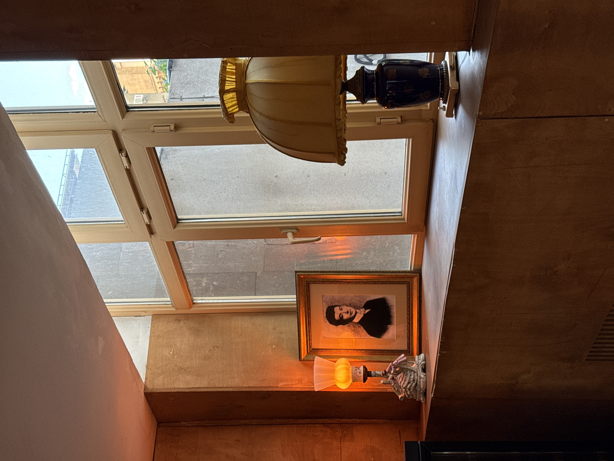 This image, when correctly oriented, provides an interior view of a room looking upwards and outwards through a window. The main subject is a large, multi-pane window with light-colored frames, likely made of wood, through which an urban outdoor environment is visible. The exterior scene includes grey building facades, a concrete or paved ground, and some green foliage, with hints of an outdoor air conditioning unit visible in one pane.
Below the window, on a wooden shelf or sill, a decorative table lamp stands prominently. It features a dark, possibly blue and gold ceramic base and a pleated, yellowish fabric lampshade. To its right, an ornate gold-framed picture displays a black and white portrait of a man, shown from the chest up, with dark hair and a dark top. Further below the picture, a smaller, ornate wall sconce is affixed to a wooden panel. This sconce emits a warm, orange-yellow light from a white, frosted glass shade, enhancing the room's cozy ambiance.
The interior walls and surfaces consist of varying materials and finishes: some areas are light-colored, possibly painted plaster, while others are paneled with wood in different shades, from natural light tones to darker stains. The lighting combines natural daylight from the window, suggesting an overcast daytime, with the warm artificial glow of the lamps. The overall impression is that of a comfortable and intimate space, possibly an attic room or a cafe. No specific activity is depicted, and no visible text is present in the image. The location context provided is Yerevan, Armenia.