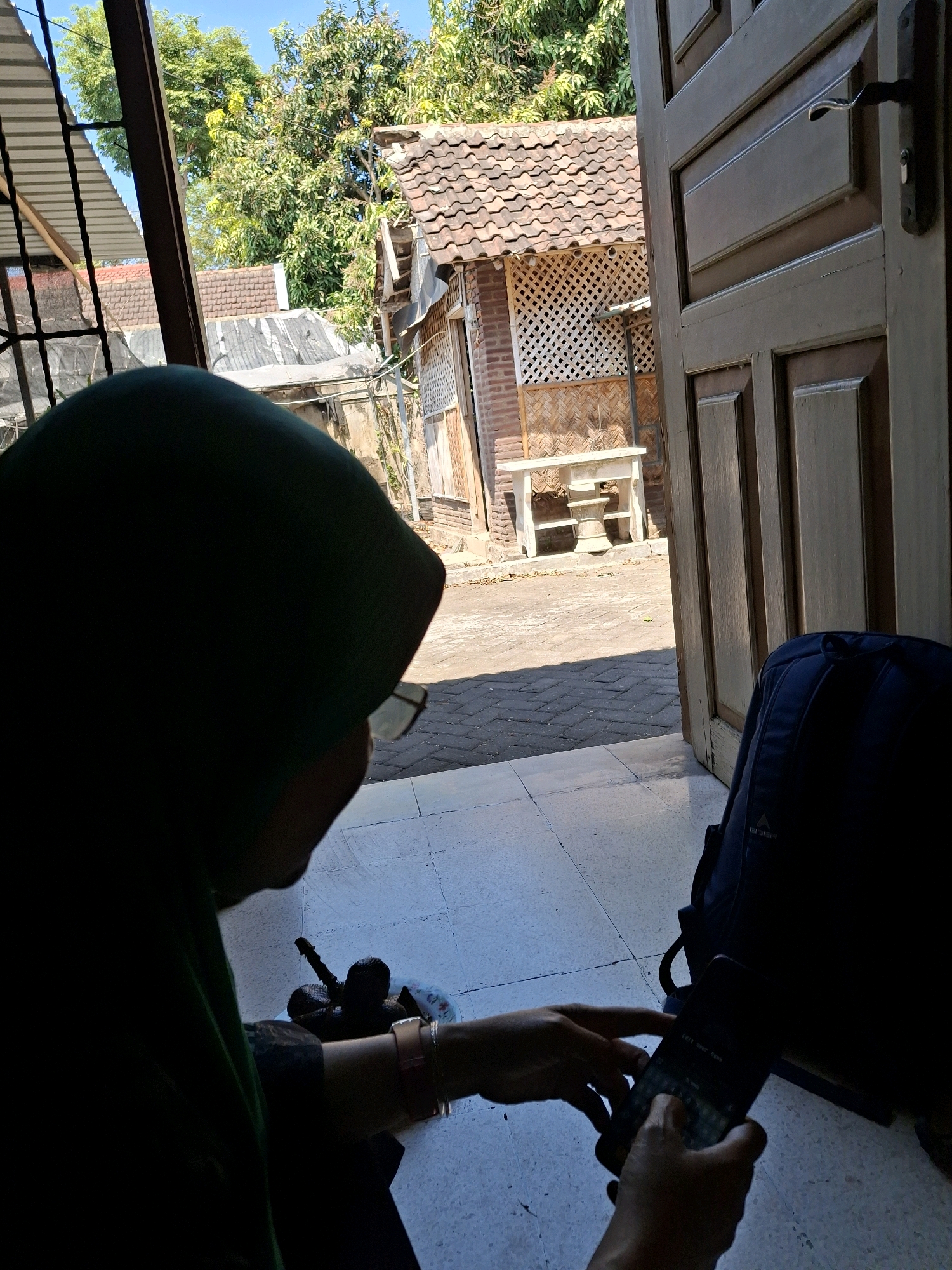 A person wearing a dark green hijab and glasses is seen from behind, seated on a tiled floor in what appears to be the entryway of a home or building. The person is holding and interacting with a smartphone. To the right of the person is a dark blue backpack. The background shows an open wooden door, revealing an outdoor scene with a tiled pathway, a low brick building with a tiled roof, and lush green trees under a clear blue sky. The environment suggests a warm, sunny day in a residential area. The style of the building and vegetation is consistent with many locations in Lumajang, Indonesia.