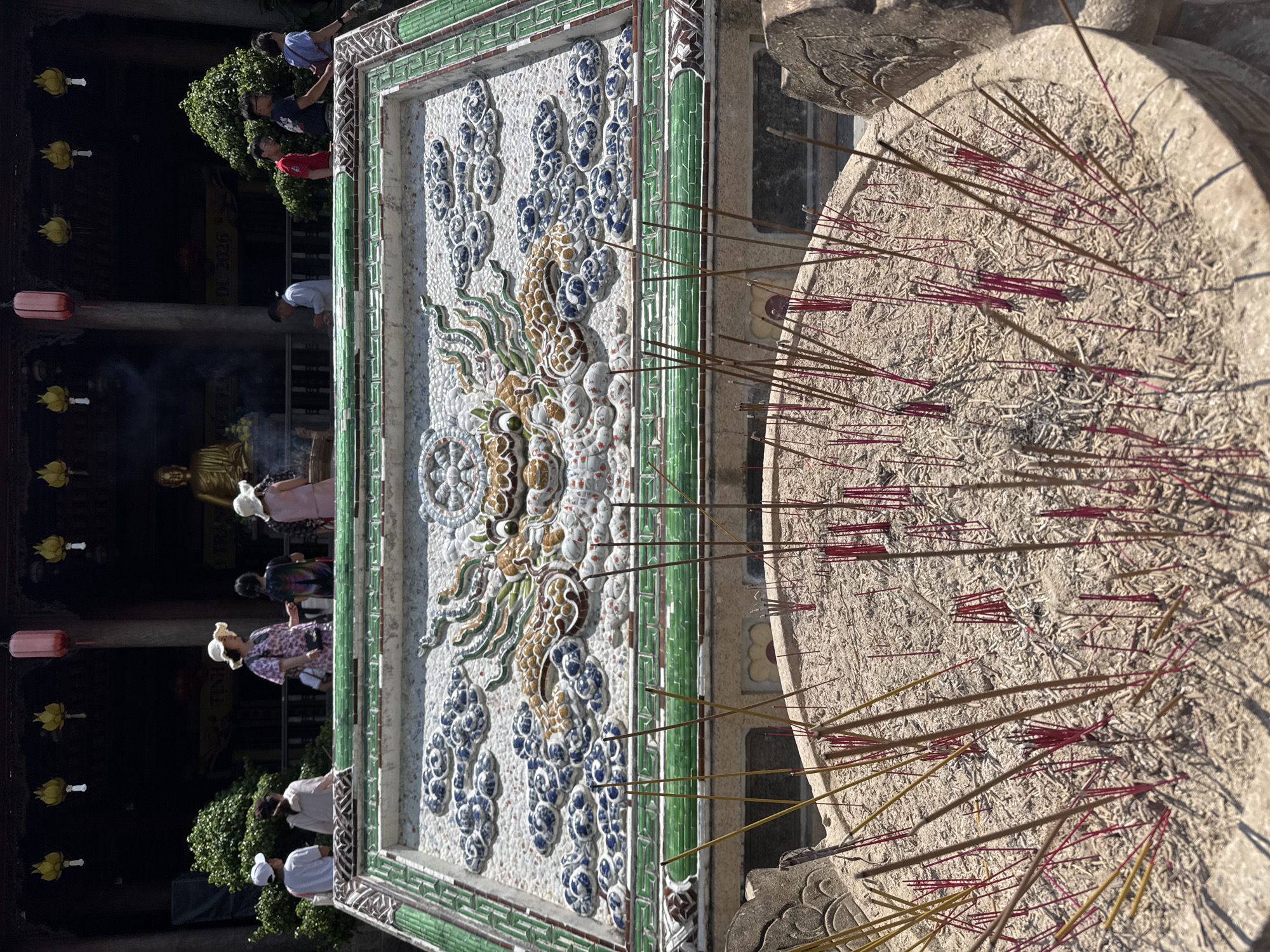 A wide shot captures an outdoor courtyard scene, likely in Tay Nha Trang Ward, Vietnam, featuring an incense burner in the foreground and a temple structure in the background. The incense burner is filled with ash and numerous lit incense sticks, predominantly red and brown, casting a subtle haze. Behind it, a large, ornate mosaic panel is visible, depicting a dragon amidst clouds. Beyond the mosaic, a temple facade is visible with people standing on a raised platform. A golden Buddha statue is visible inside the temple, and red and yellow lanterns hang from the eaves. Several individuals are present on the temple's upper level, some standing and observing, while others appear to be interacting. The overall environment suggests a place of worship or cultural significance.