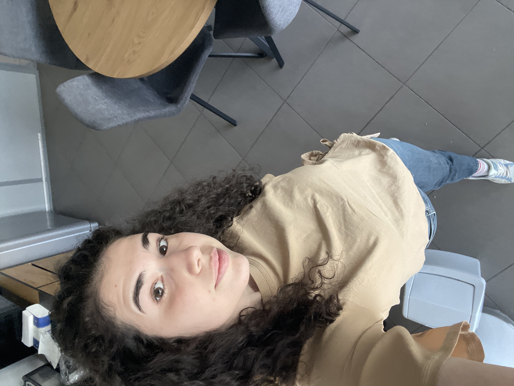 A woman with dark, curly hair is pictured in a medium-shot selfie, looking at the camera. She wears a tan-colored top and blue jeans and the leg stretches out to the right of the frame to reveal tennis shoes. The ground is a gray tile. In the background are two gray chairs and a round table. In the left of the frame is a white appliance that is likely a refrigerator or ice maker. This image was taken in Sofia, Bulgaria.