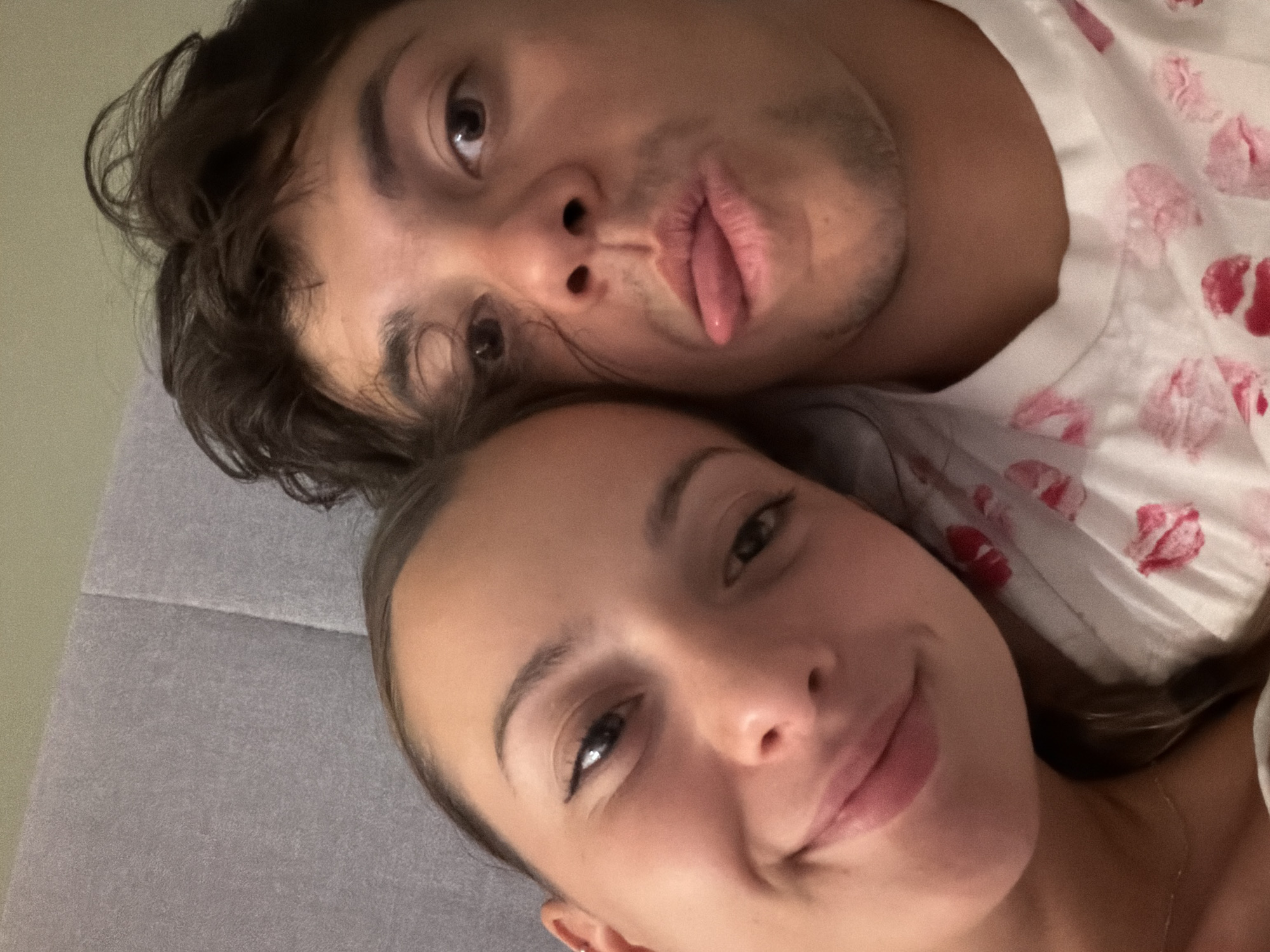 This is a close-up, selfie-style photograph of a young couple in Sofia, Bulgaria, lying down, likely in bed, as indicated by the gray textured headboard and the soft lighting. The woman is on the left, smiling at the camera, with her left ear visible and a subtle piercing. She has dark eyeliner and a light foundation. The man is on the right, making a playful expression with his tongue sticking out slightly. He has dark, messy hair and stubble on his face. Both are wearing t-shirts; the man's is white with a pattern of red lipstick kiss marks. The background is a muted green wall.