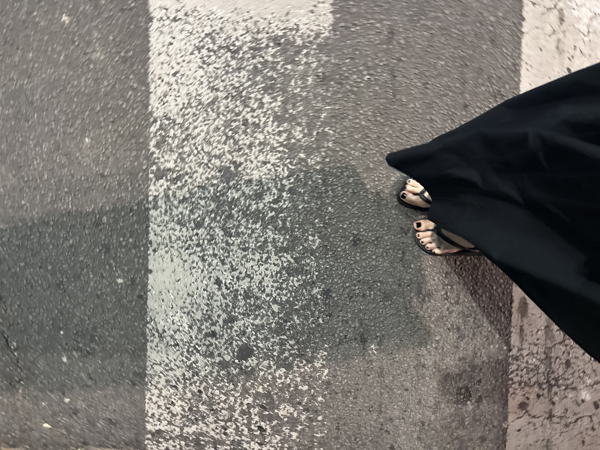 The image depicts a pedestrian standing on a zebra crossing in Vracar Urban Municipality, Serbia. The crossing consists of alternating stripes of light and dark grey concrete. The pedestrian's feet, adorned with black nail polish, are visible in black flip-flops. A portion of a black garment or fabric obscures part of the scene on the right side of the image. The concrete surface shows signs of wear and discoloration.
