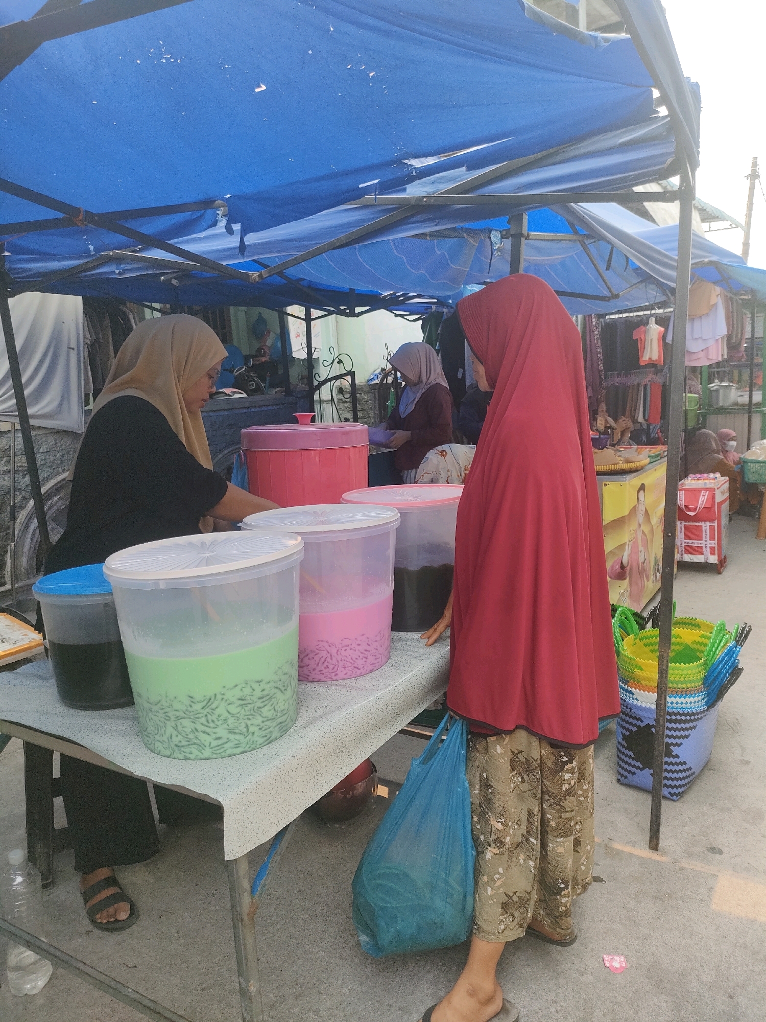 A street food vendor operates a stall under a blue tarp in Batam, Indonesia. The vendor, wearing a red hijab and patterned pants, stands facing away from the camera, looking towards a large translucent container filled with a green, jelly-like dessert and dark liquid. To the left, another vendor in a light brown hijab, wearing a black shirt and dark pants, serves customers. In front of the vendors are several large plastic containers of drinks and desserts. The stall is set up on a table covered with a patterned cloth. Behind the stall, clothing racks and other items suggest a marketplace setting. The ground is paved, and a blue plastic bag is visible on the right side of the image.