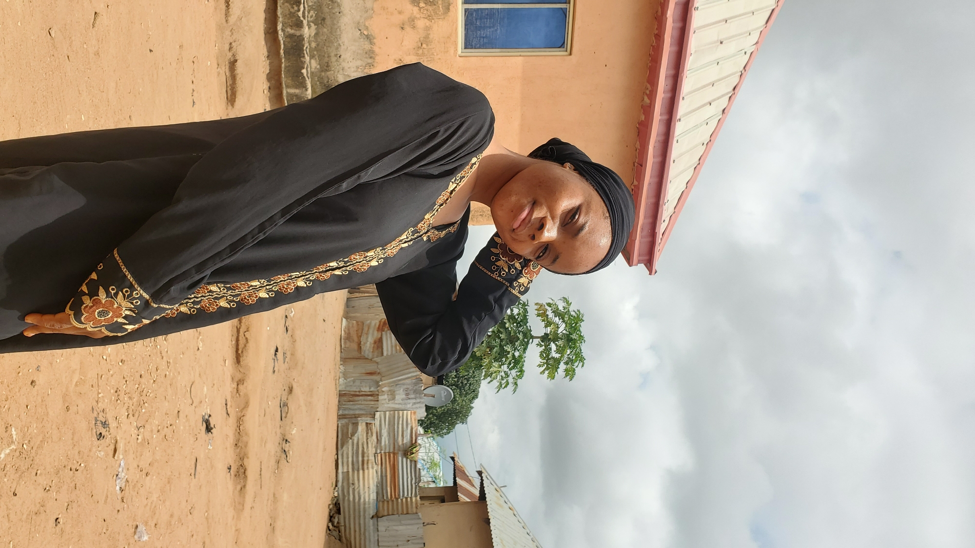 A young woman wearing a black abaya with gold embroidery and a black headscarf is posing for a portrait outdoors in Jalingo, Nigeria. She is standing in front of a peach-colored building with a corrugated metal roof and a blue-paned window. Her left arm is bent, with her hand resting on the back of her head, and she is looking at the camera with a slight smile. The background shows a dusty, unpaved area with corrugated metal fences, a small tree, and another building with a metal roof. The sky is cloudy.