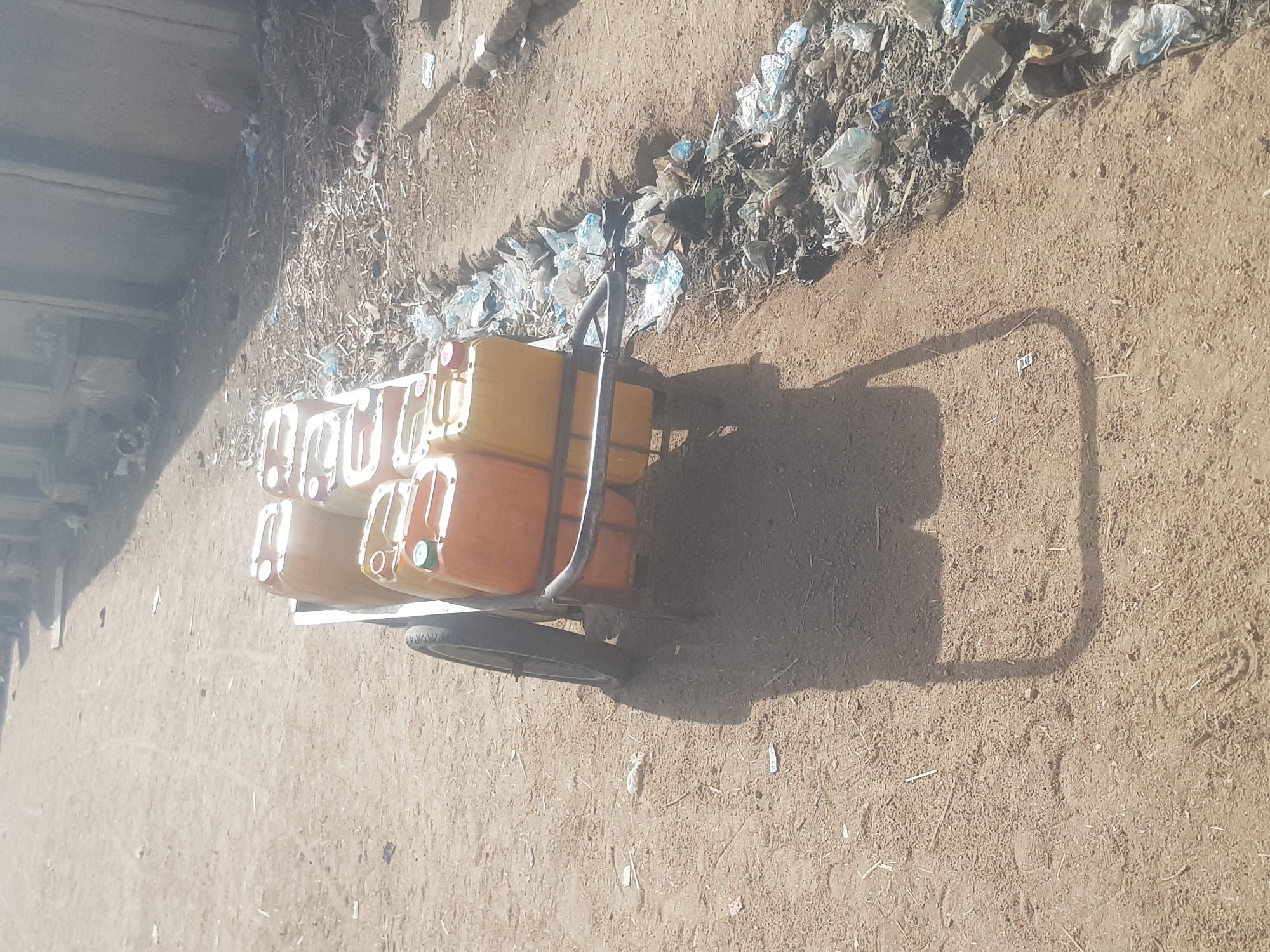 In Dutsinma B, Nigeria, a two-wheeled cart loaded with approximately ten yellow jerrycans sits on a dirt surface. The cart is positioned near a trash-filled ditch. The environment appears arid, and shadows indicate a sunny day. There are no people visible in the immediate frame.