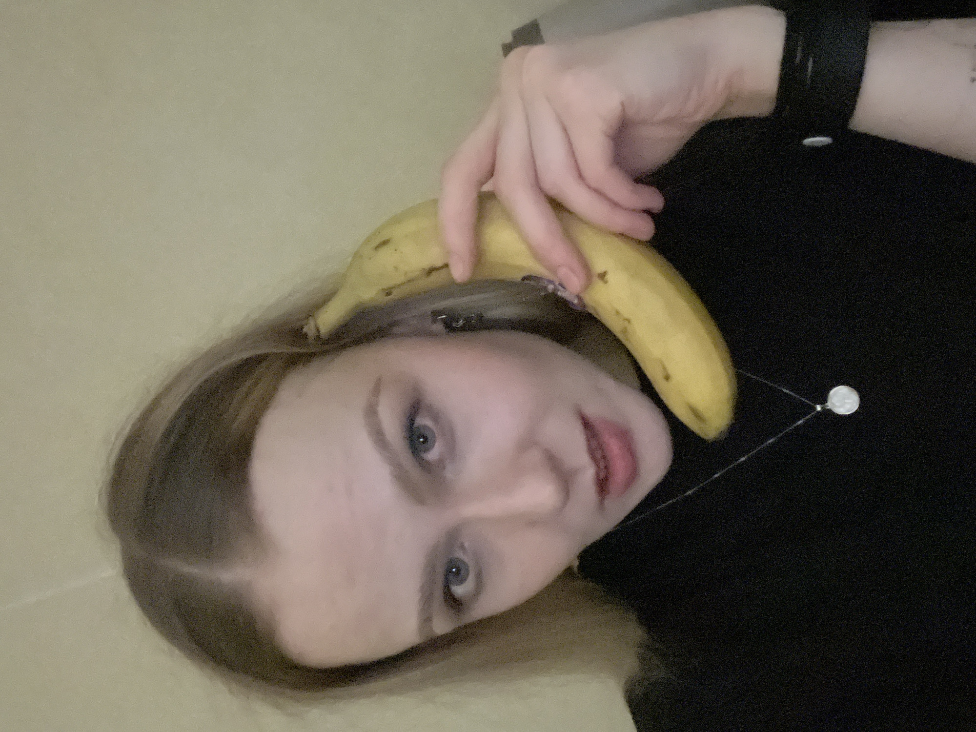 A young woman with blonde hair is holding a yellow banana near her ear, seemingly mimicking a phone. She is wearing a black shirt and a black watch. A silver necklace with a round pendant is visible. The background appears to be a light beige wall. It is not possible to determine the specific location in Khimki, Russia from the image.