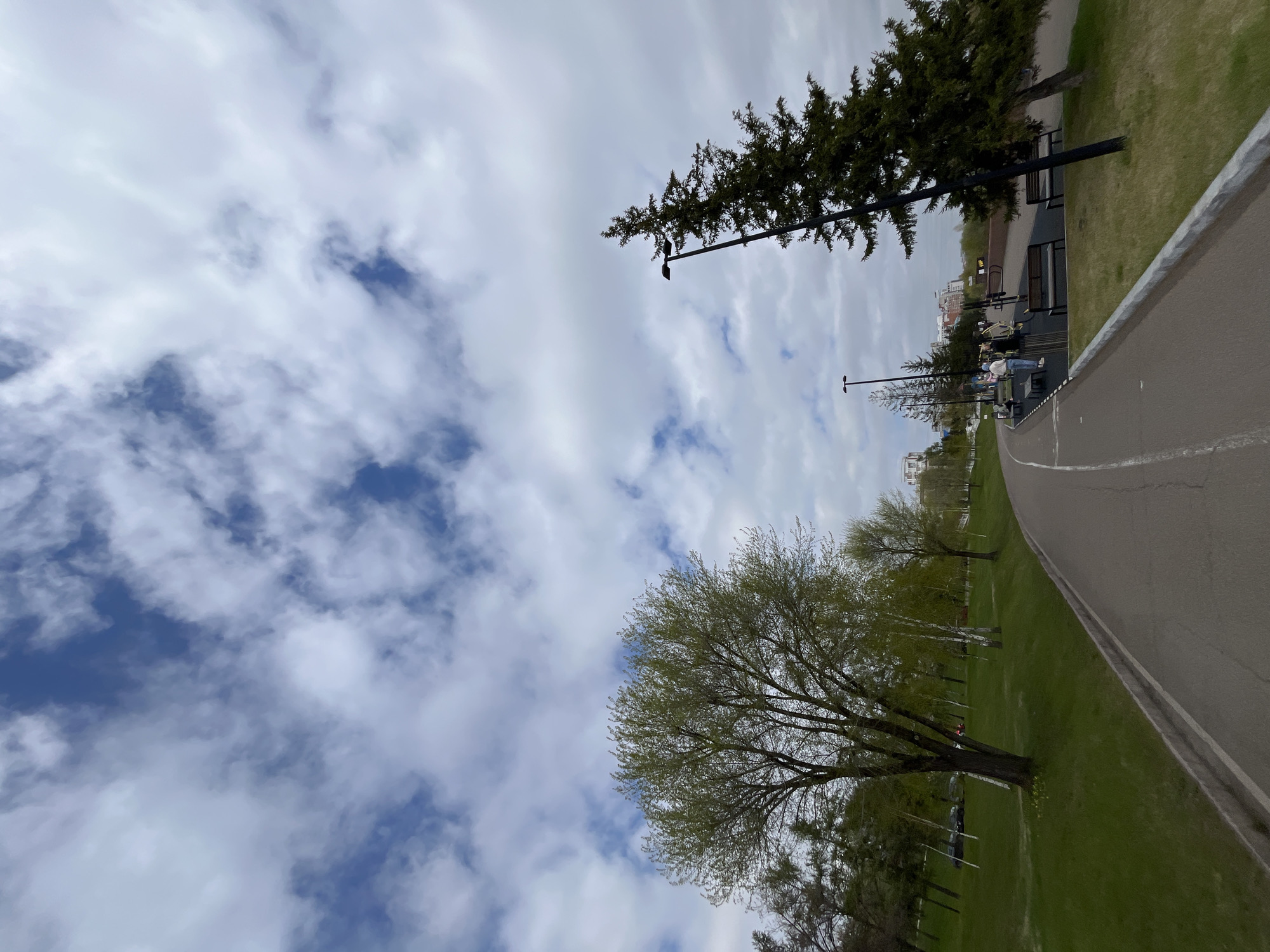 The image shows a view of a park on Tatyshev Island in Krasnoyarsk, Russia. The scene is tilted, with the sky featuring white clouds and patches of blue. A paved path curves along the right side of the image, bordered by green grass. Trees line the path, including a tall evergreen and a tree with fresh spring leaves. Benches and workout equipment are visible along the path, with several people using the equipment. Streetlights are positioned along the path. Buildings can be seen in the background.