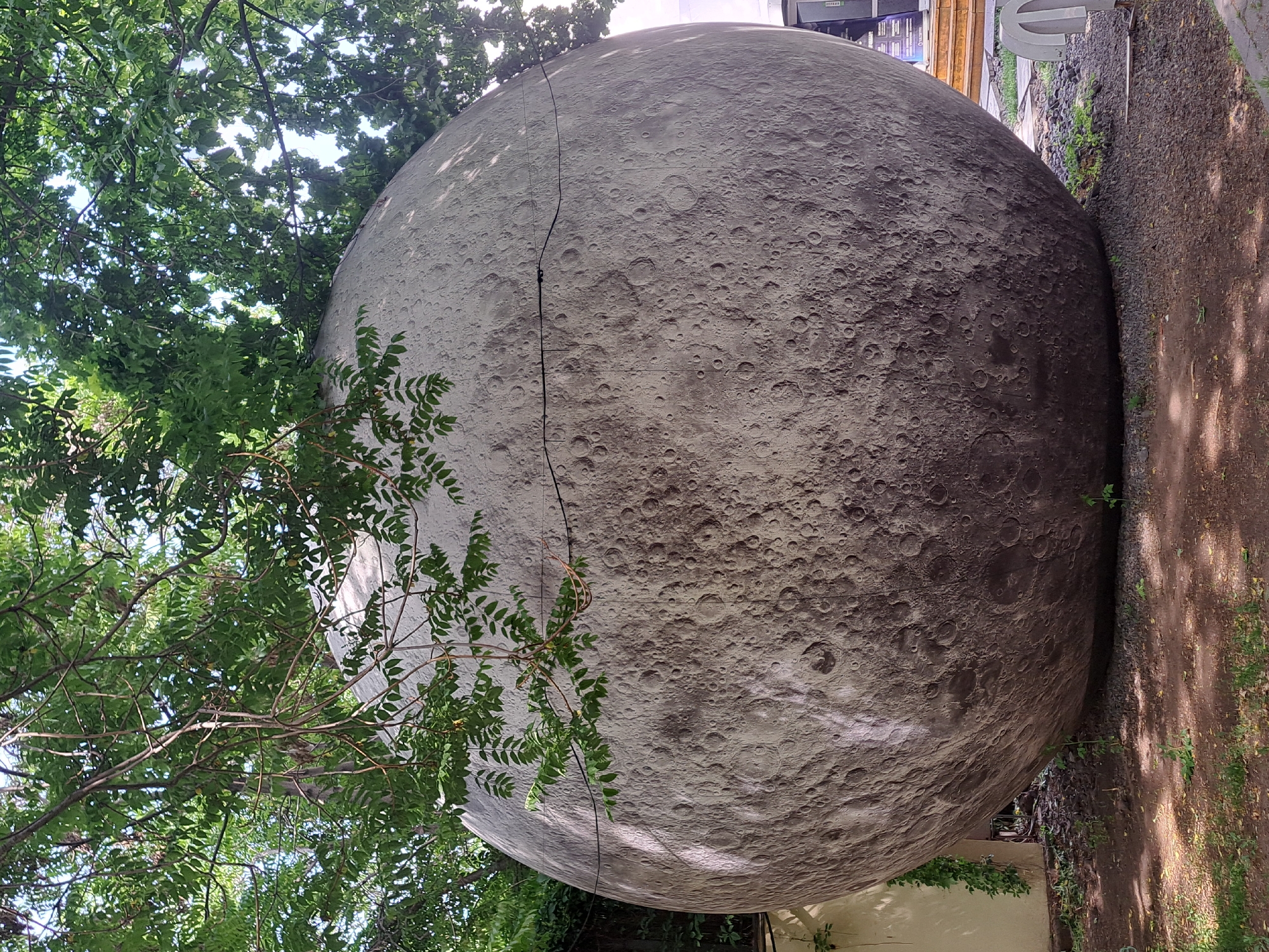 The image depicts a large-scale model of the moon, its surface detailed with craters and varying shades of gray, situated outdoors. The structure appears partially obscured by foliage from a tree on the left. A thin, dark cable runs vertically across the moon's surface. The moon replica is positioned next to a path or area covered with fine gravel or small stones. Based on research, this is the Yerevan Moon, located in the city of Yerevan, Armenia.