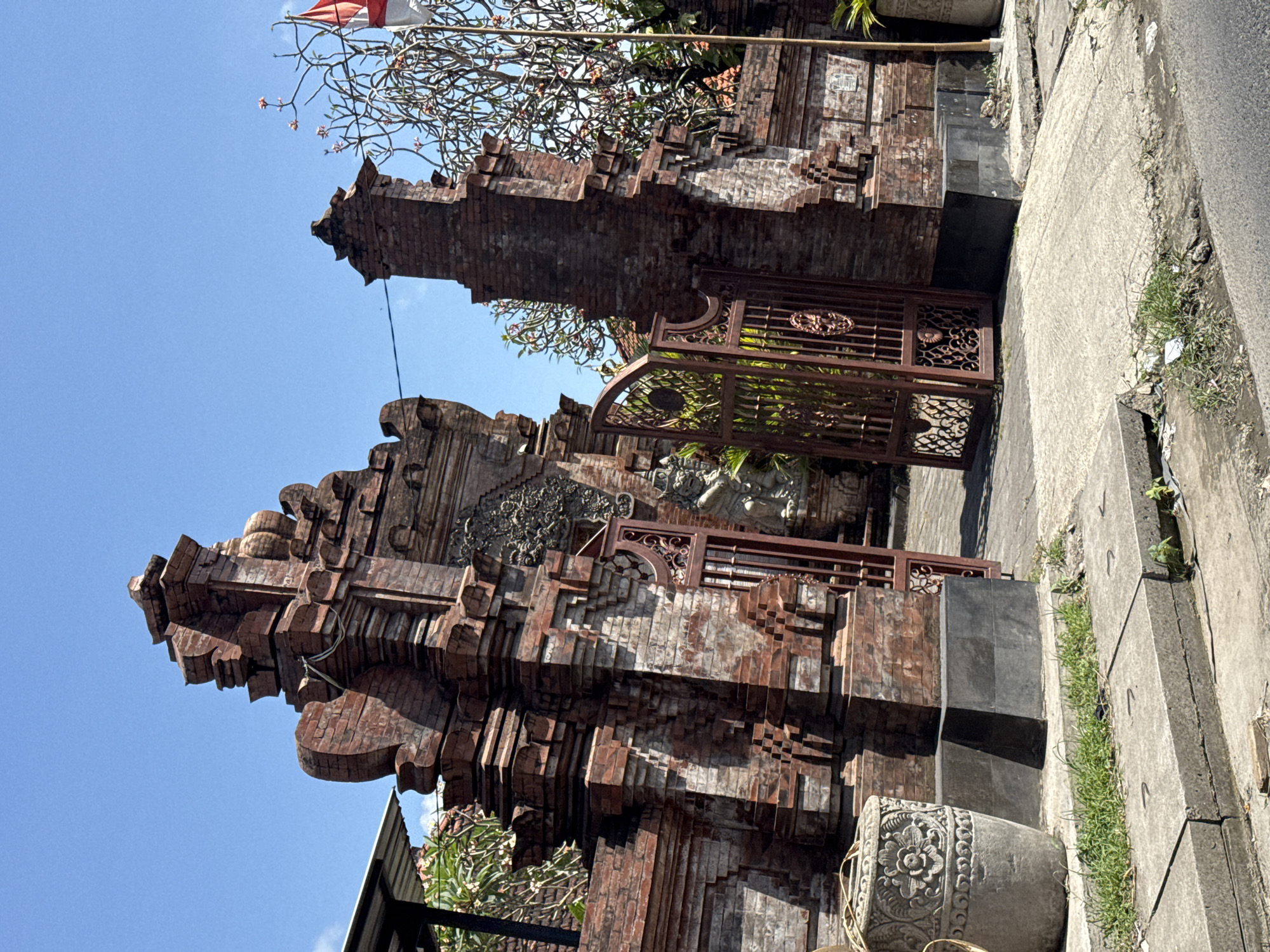 An ornate brick gate, likely a temple entrance, stands under a clear blue sky in Denpasar, Indonesia. The gate features intricate carvings and decorative elements typical of Balinese architecture. A metal gate, also ornate, is partially open, revealing greenery within. A large, carved stone pot sits to the left of the gate. A flagpole with a red and white flag is visible to the right, and a frangipani tree with small pink flowers is also present. The gate is constructed from weathered red brick and appears to be a significant architectural feature. The ground in front of the gate is paved with concrete and shows signs of wear. The overall scene suggests a warm, sunny day in a tropical location.