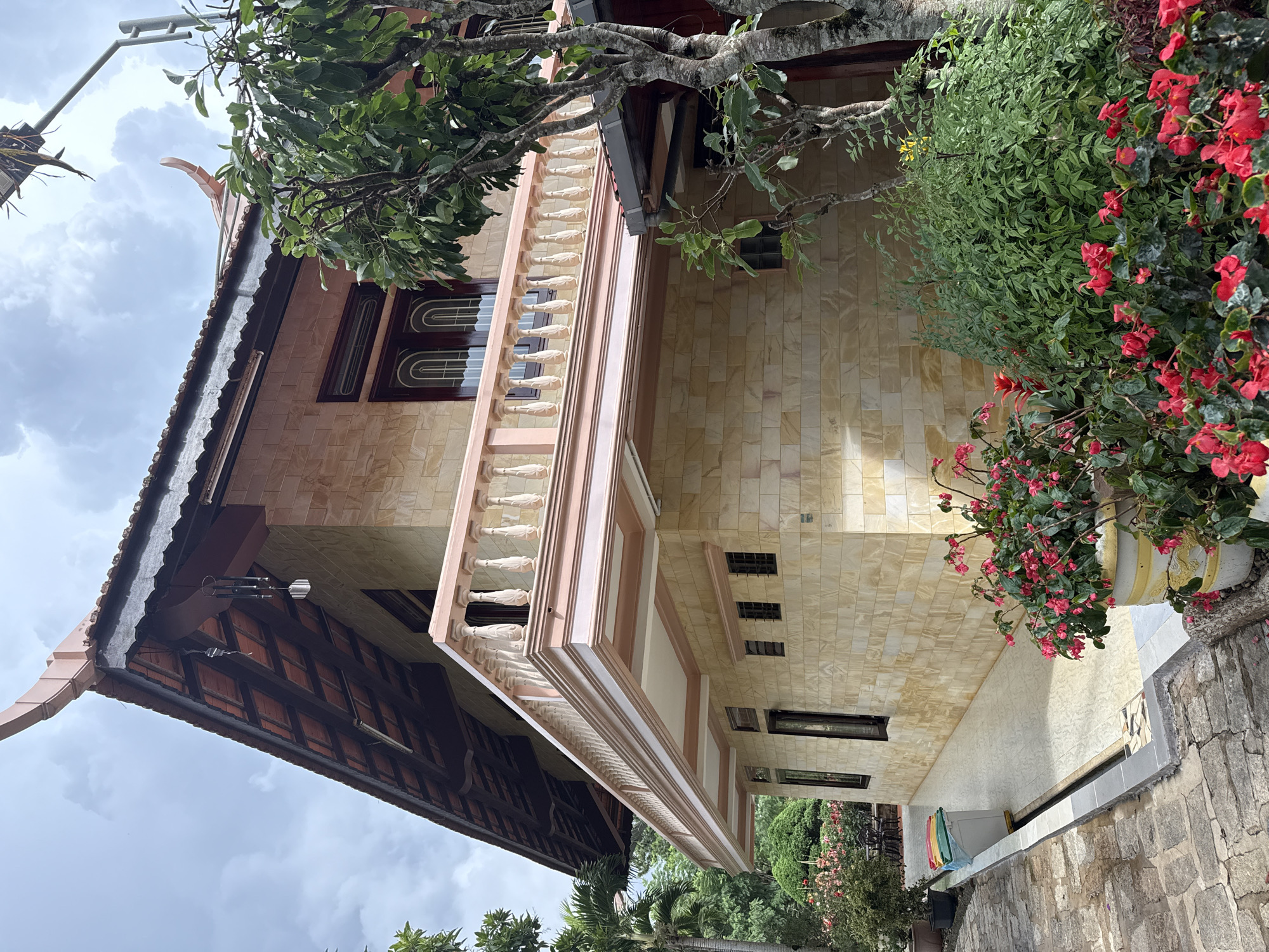 The image depicts the exterior of a two-story building, likely a house or a guesthouse, in the city of Da Lat, Vietnam. The architecture features a distinct tiled roof with curved eaves and a balcony adorned with ornate balusters. The building's facade is constructed from light-colored, rectangular stone tiles. In the foreground, a pathway paved with cobblestones leads towards the building, bordered by lush greenery and vibrant red flowers in planters. The surrounding vegetation suggests a garden setting, common in Da Lat's cooler climate. The sky is overcast with a few scattered clouds, indicating a diffused daylight. No people are visible in the scene. The overall impression is one of a well-maintained property with a blend of traditional architectural elements and natural landscaping.