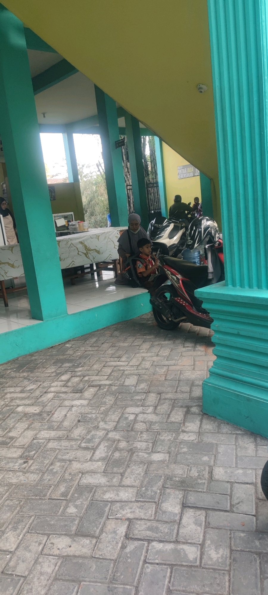 In Banjarsari, Indonesia, a child wearing a scout uniform sits on the seat of a red motorcycle, facing left. Beside the child, a woman in a grey hijab and robe sits on the same motorcycle, also facing left. The motorcycle is parked on a brick-patterned pavement. In the background, there are several other motorcycles, including a white one with two people on it. A table with a patterned cloth is visible behind the people on the motorcycles, with another person standing behind it. The scene appears to be an outdoor area with teal-colored columns and a yellow ceiling structure. A sign with text is also visible on the yellow structure in the background.