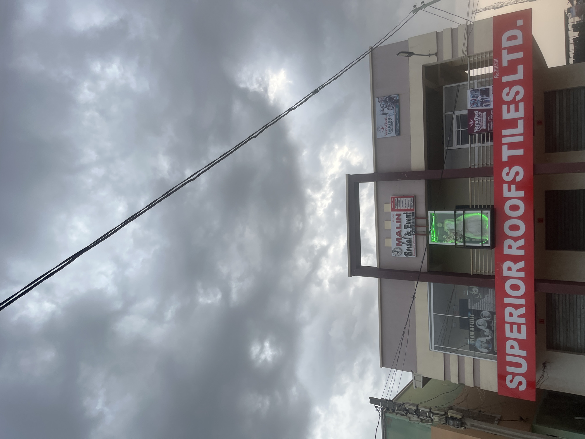 The image shows a commercial building with a large red sign that reads "SUPERIOR ROOFS TILES LTD." The building has multiple floors and several businesses are advertised on the upper floors. One sign reads "MALIN Bridal & Event," and another displays "Verified Baking Academy." A neon green sign depicts a person in a dress, likely a wedding gown. The sky is overcast with dramatic, dark clouds. Power lines are visible in the foreground, stretching across the frame. The building appears to be located in an urban or semi-urban area, possibly Gura Top, Nigeria, given the context.