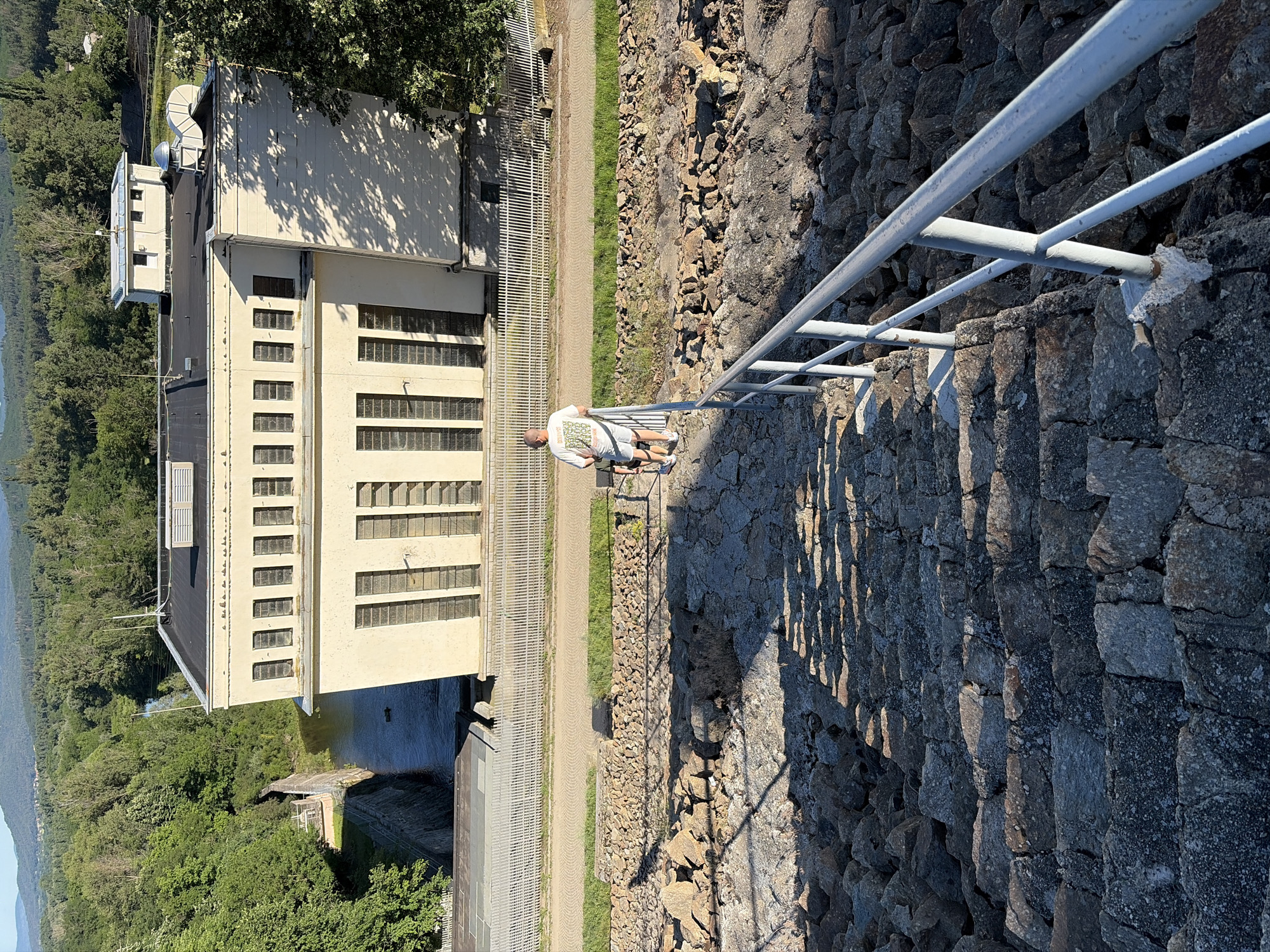 The image shows a man standing on a stone wall next to a building. The man is wearing a white t-shirt with a graphic design and gray shorts. He is leaning on a light-colored metal railing. The building appears to be a power station or dam, with a large concrete structure and numerous windows. A body of water is visible behind the building, and a lush green forest surrounds the area. The sun is casting long shadows, suggesting it is either early morning or late afternoon. The weather appears to be clear and sunny. The setting is outdoors, possibly at a hydroelectric dam in Koprinka, Bulgaria. The overall mood is peaceful and scenic. There is no visible text in the image.