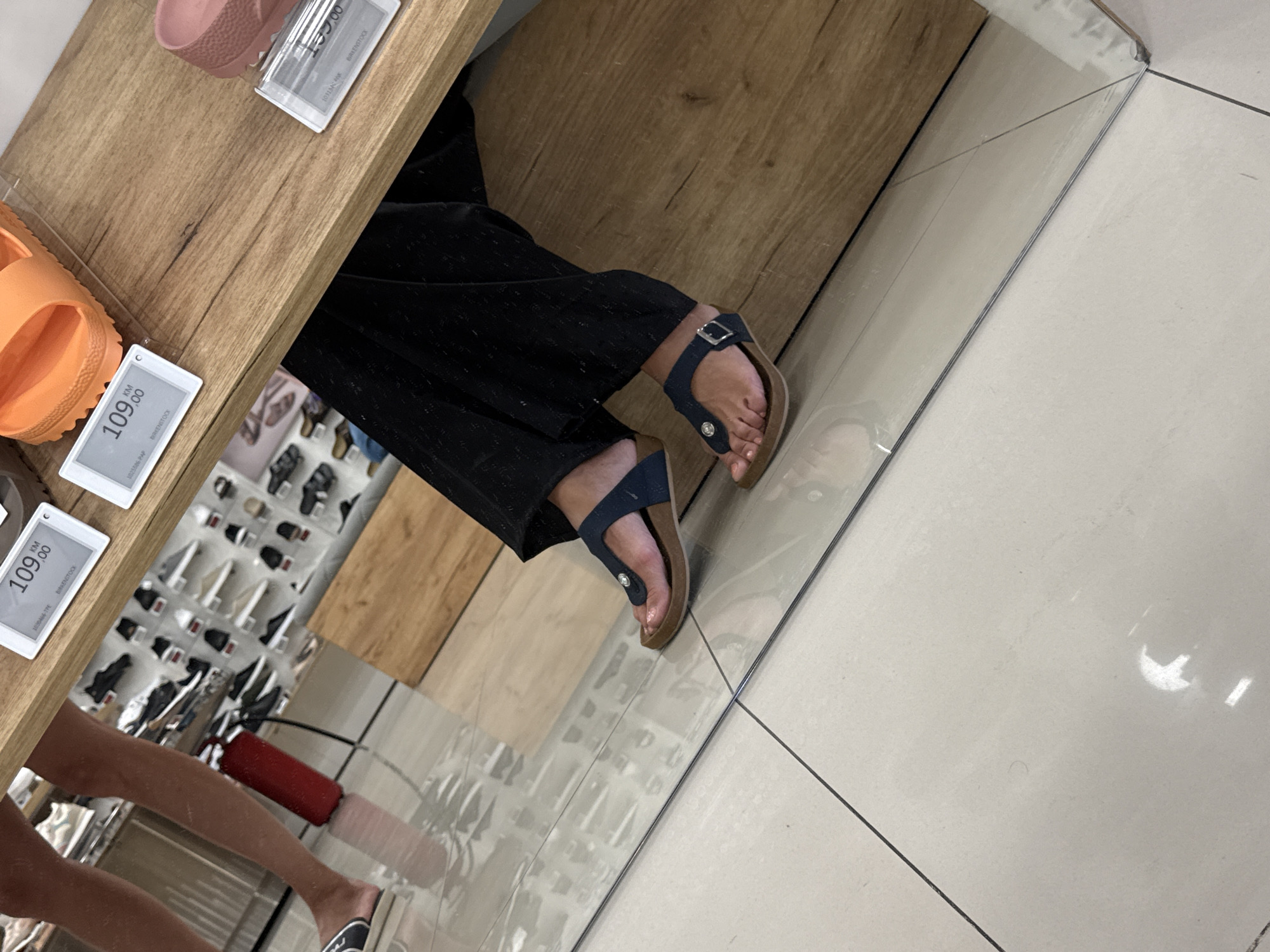 The image depicts a close-up view of a person's feet and legs, wearing dark pants and dark blue thong sandals, standing on a tiled floor. The person is positioned in what appears to be a retail environment, possibly a shoe store, as evidenced by display shelves filled with footwear visible in the background and price tags on a wooden surface in the foreground. One price tag displays "109,00 KM" and "BIRKENSTOCK". The floor is composed of large, light-colored tiles with dark grout lines. A reflection of the environment is visible on the floor. The scene is illuminated with artificial lighting, casting reflections and highlights. The location is identified as Ilidža, Bosnia and Herzegovina, suggesting this is a retail establishment within that city.