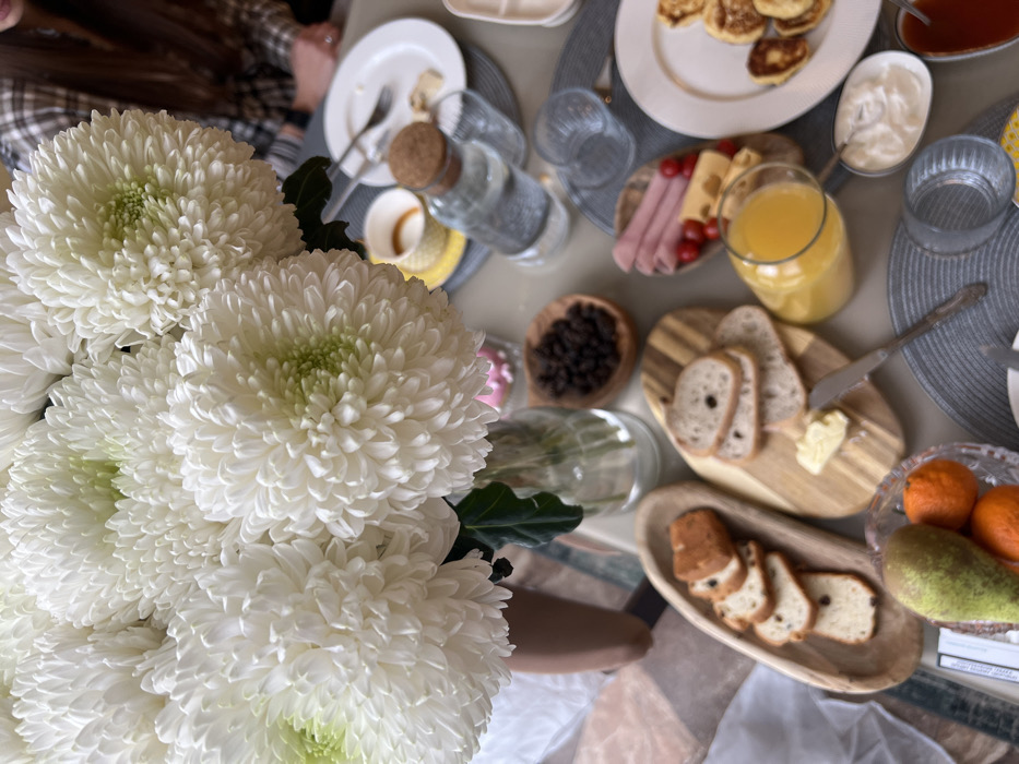 The image depicts an indoor breakfast or brunch scene on a light-colored table in Khimki, Russia. A large bouquet of dense white chrysanthemums occupies the left foreground in a clear glass vase. On the table, a variety of food items are presented: slices of fruit bread in a wooden oval bowl, round bread slices with butter and a silver knife on a wooden cutting board, ham, cheese cubes, and cherry tomatoes in a small wooden tray, and several pancakes on a white plate. A small white bowl contains a creamy substance, likely yogurt or sour cream. Beverages include orange juice in a clear jug, water in a cork-stoppered glass bottle, and a partially consumed drink in a small yellow and white teacup. A crystal bowl in the lower left holds a green pear and two tangerines. Several clear drinking glasses, white ceramic plates, silverware, and gray woven placemats are arranged on the table. A person with a plaid shirt and a ring on their right hand is partially visible, out of focus, in the upper right background.