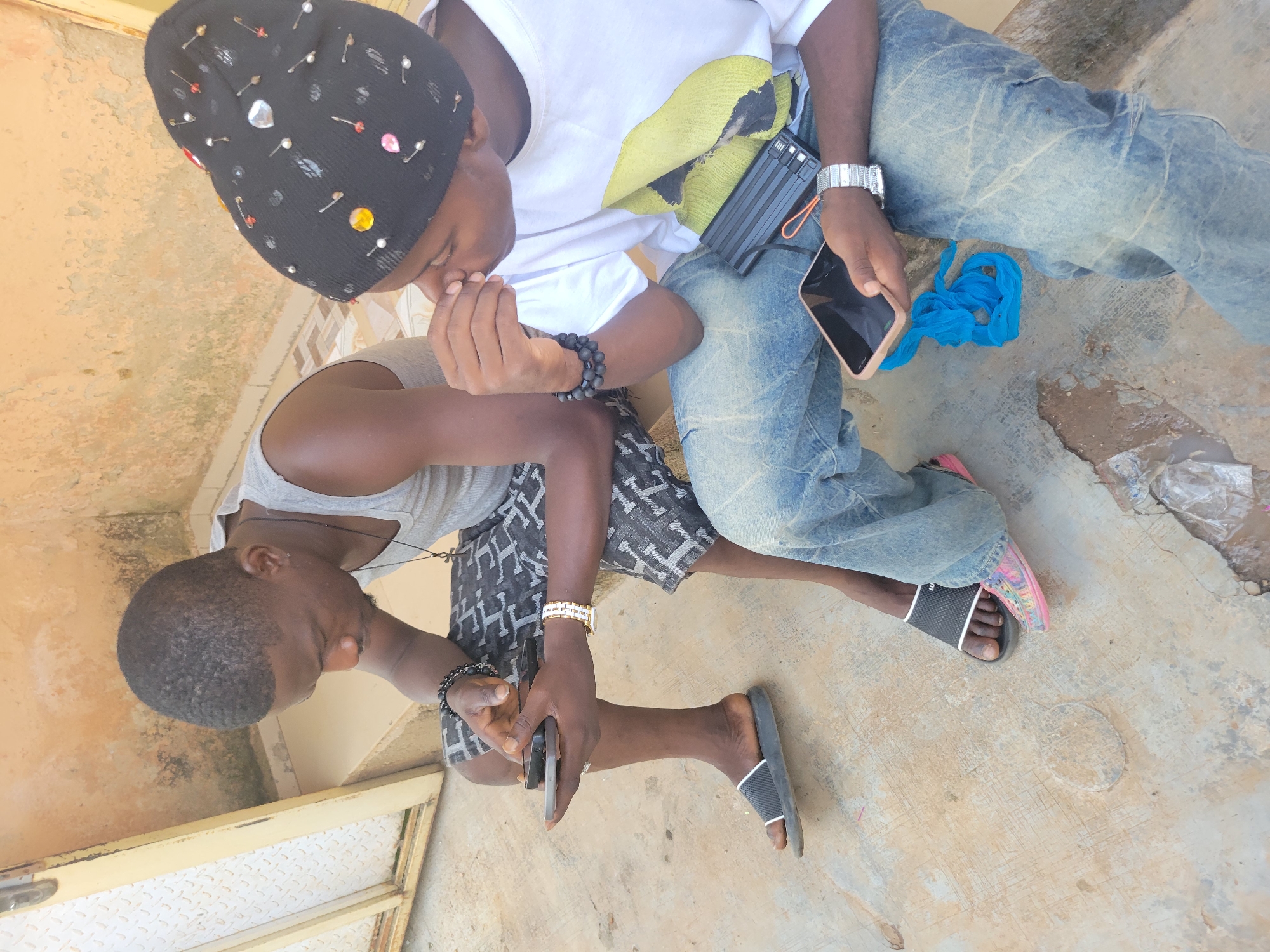 Two young men are sitting outdoors on a concrete surface next to a building with textured, orange-colored walls. One man, on the left, wears a grey tank top and patterned shorts, with black beaded bracelets on his left wrist and a gold watch on his right. He is holding two mobile phones. The other man, on the right, wears a black beanie adorned with colorful beads and pins, a white t-shirt with a graphic print, and distressed blue jeans. He has black beaded bracelets on his left wrist and a silver watch on his right. He is holding a mobile phone and there is a black power bank and a blue plastic bag next to him. Both men are looking down at their phones, appearing engaged with their devices. The ground is stained and appears worn, with some debris scattered around. The environment suggests a casual, urban setting.