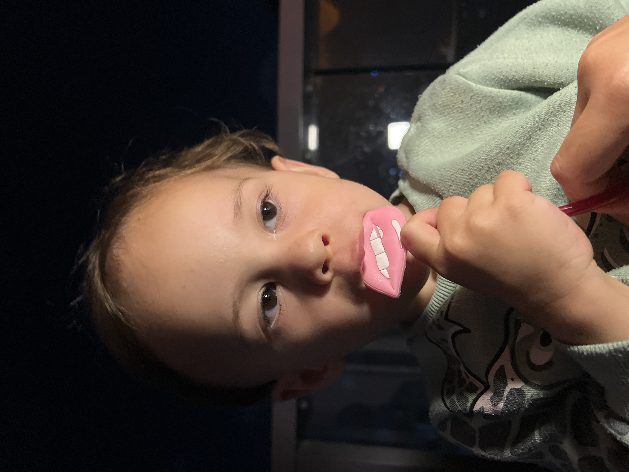 A young child with light brown hair and eyes is turned at an angle, looking directly at the camera. The child is holding a pink plastic toy with white tooth shapes in their mouth. Their other hand holds a red object, possibly a toothbrush. The child is wearing a light green sweatshirt with a cartoon-like print on the sleeve. The background is dark, but a window is visible with lights visible through it. No specific location is deducible from the image. The mention of Kiryat Bialik, Israel, is not possible to verify with the image alone.