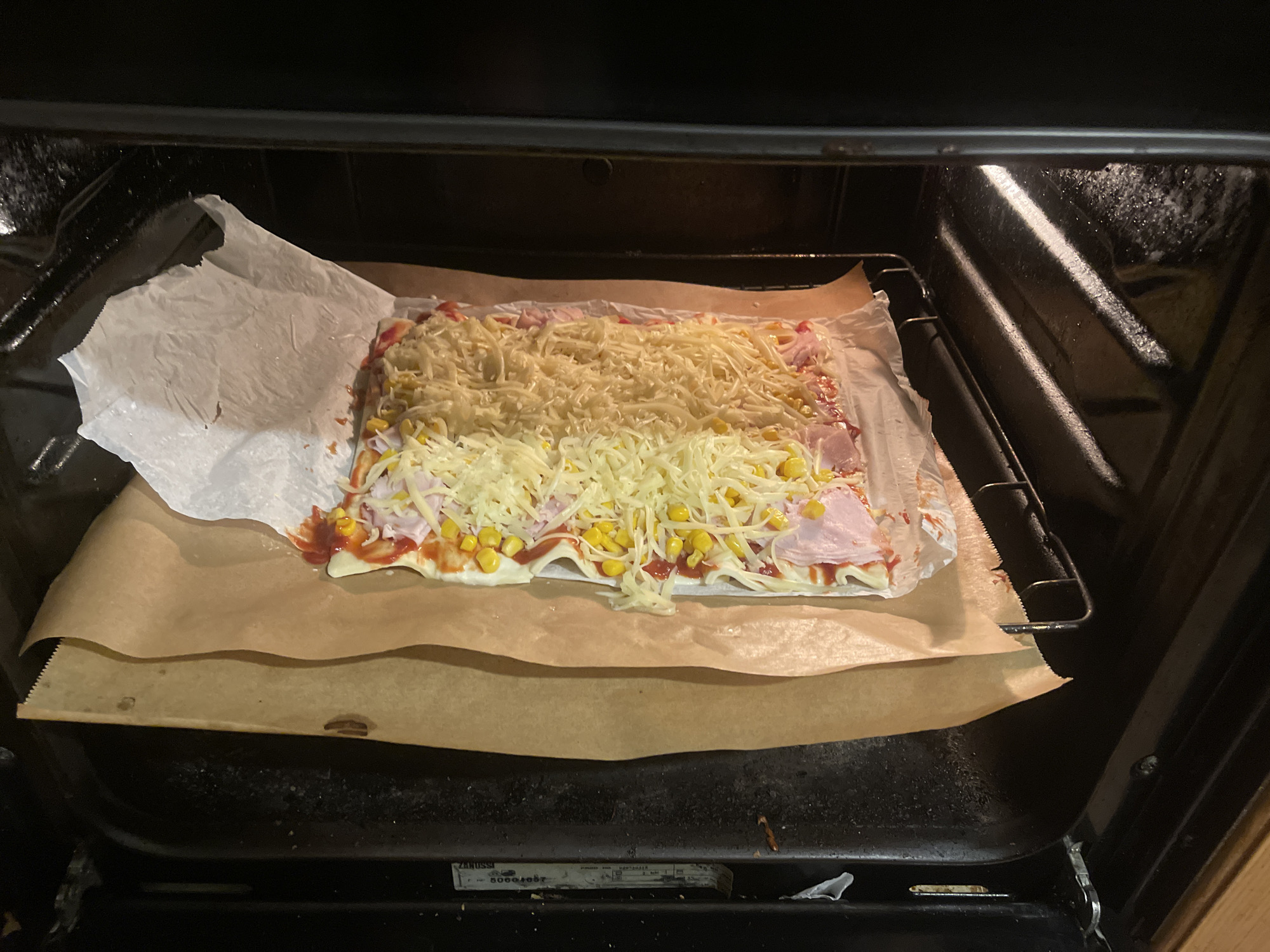 A pizza sits on parchment paper inside an oven. The pizza is topped with tomato sauce, ham, corn, and shredded cheese. A piece of wax paper lies next to the pizza. The oven's interior is dark. The location is Unknown, Unknown.