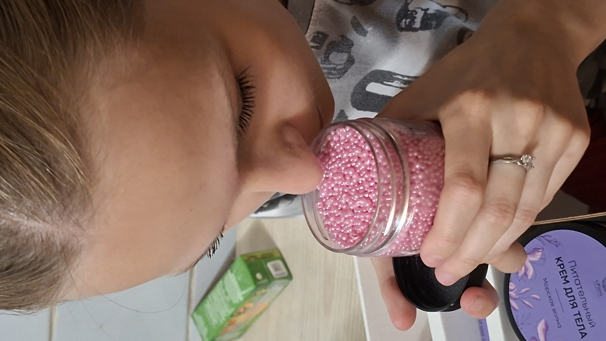A close-up shot shows a person's face from the side as they lean in to smell a small, clear jar filled with pink beads. The person has blonde hair and long eyelashes. Their eyes are closed, suggesting they are focusing on the scent. The hand holding the jar is adorned with a ring on the ring finger, featuring a small, clear stone. In the background, a green box and a purple container with Cyrillic text are visible. The purple container has text that reads "ПИТАТЕЛЬНЫЙ КРЕМ ДЛЯ ТЕЛА" and "Морской волна" which translates to "NOURISHING BODY CREAM" and "Sea wave". The image is well-lit, indicating it was taken indoors during the daytime. The overall mood appears to be one of curiosity and sensory exploration.