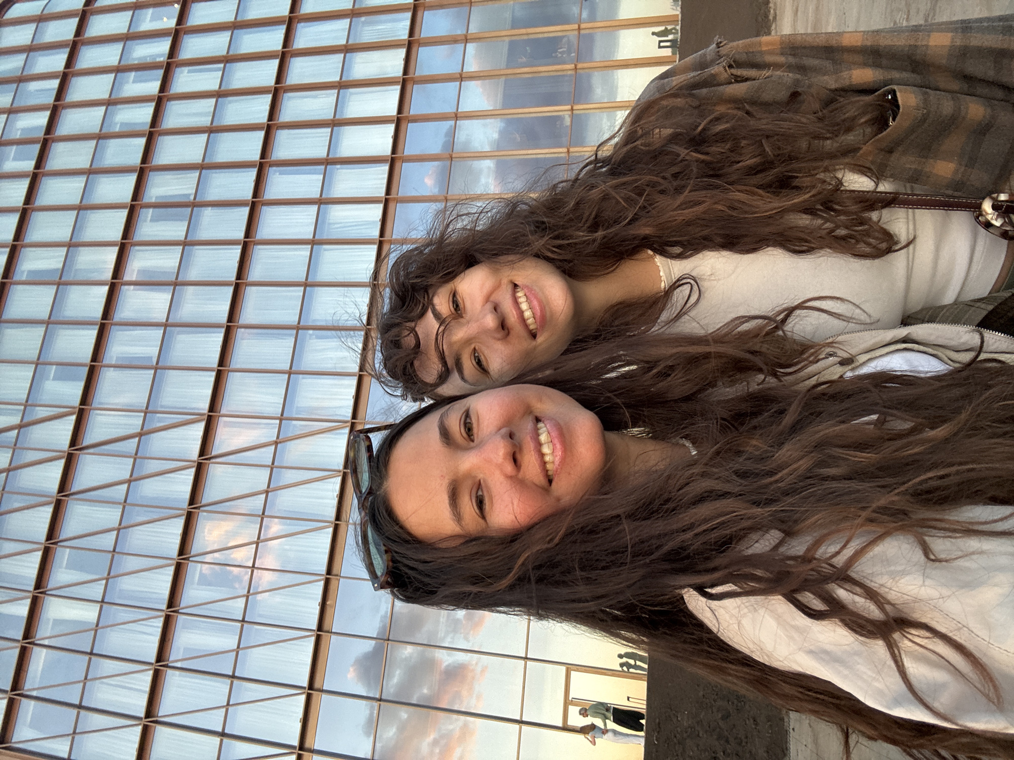 Two young adult females are captured in a close-up, outdoor selfie, smiling directly at the camera. The woman on the left has long, wavy dark brown hair, wearing a light-colored jacket over a white top, with dark sunglasses resting on her head. Her face shows a broad smile with visible teeth. The woman on the right has long, curly dark brown hair, also smiling broadly with visible teeth. She wears a light-colored top beneath a plaid brown and tan jacket, and a dark strap crosses her chest.
The background is dominated by a multi-story modern building with a geometric facade. The building's exterior features a grid of reddish-brown or bronze-colored framing around numerous rectangular glass windows. Some sections of the facade, particularly on the left, incorporate diagonal bracing within the rectangular panes. The windows strongly reflect the sky and clouds, showing warm hues of pink, orange, and white clouds against a pale blue sky, indicative of either sunset or sunrise lighting. In the reflections on the lower-left portion of the building, several small, indistinct human figures are visible, including what appears to be a couple. The immediate foreground where the subjects stand is a dark, flat, possibly unpaved surface. The location is described as a city, specifically Kazan, Russia.
