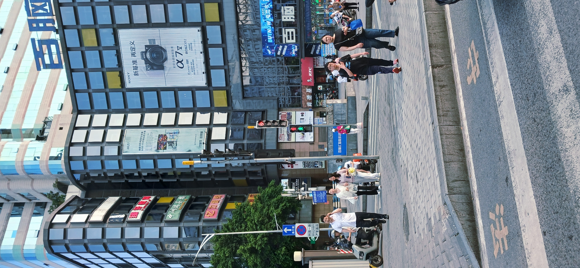 The image is of a street scene in Jiulongpo District, China. The shot focuses on the people and buildings alongside the road.
**People:** A group of pedestrians are walking on the sidewalk and crosswalk. A man and a woman are walking toward the camera. Another group of people is walking away from the camera.
**Buildings:** Modern buildings with large advertising signs are visible. Some of the signs advertise brands like Sony, KFC, and 百脑汇 (Buynow).
**Objects:** Traffic lights, street signs, and parked vehicles are also present.