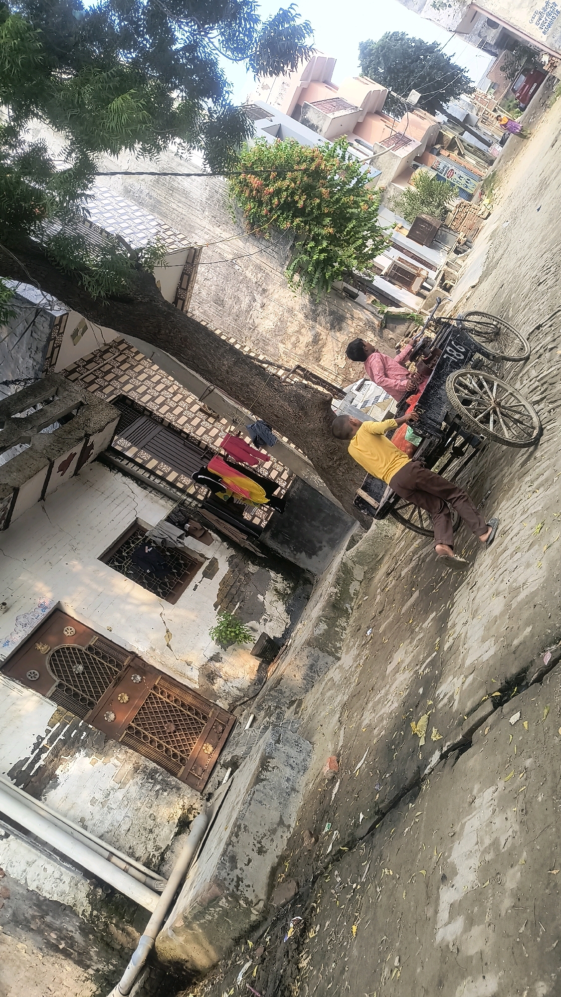 The image depicts an outdoor scene in Loni, India, characterized by a narrow, unpaved street lined with buildings. In the foreground, a large tree with dense green foliage extends into the frame from the left, its trunk partially obscuring a white-washed building. This building features a decorative brown metal gate and a barred window. Adjacent to the tree, clothing is hung on a rack, including a pink shirt, a yellow garment, and dark trousers.
Further down the street, a black cart with two large spoked wheels is visible. Two individuals are interacting with the cart. One person, wearing a yellow shirt and brown trousers, is leaning against the cart. Another person, wearing a pink shirt, is seated inside the cart. The street itself is uneven and appears to be made of packed dirt and concrete, with debris scattered across its surface. In the background, several buildings of varying architectural styles and colors are visible, suggesting a densely populated urban environment. The sky above is partly cloudy, with sunlight casting shadows across the scene.